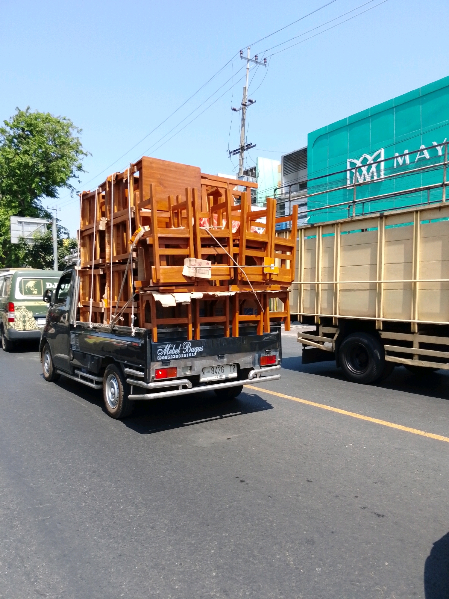 In Pasuruan, Indonesia, on a clear, sunny day, a black pickup truck loaded with wooden chairs is traveling on a road. The chairs are stacked high, some reaching above the cab of the truck. The truck has a license plate that reads "N 8426 TO". To the right of the pickup truck, a larger truck with a turquoise colored cargo box is also on the road. The cargo box has white lettering that appears to say "MM MAY". The environment suggests a commercial area or a road within the city, with buildings and trees visible in the background. The sky is a vibrant blue.