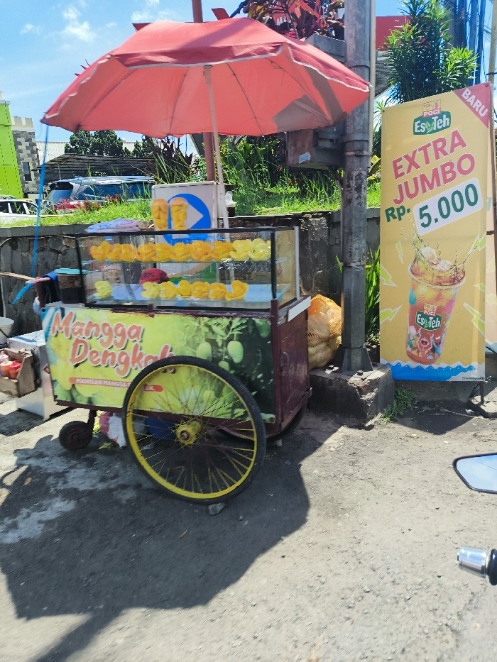 The scene is a street corner in Padalarang, Indonesia. A street vendor has set up a cart selling fruit, specifically mangoes, as indicated by the sign "Mangga Dengka." The cart is equipped with a red umbrella for shade. The cart is parked against a light pole, with a large advertising sign for a drink called "Es Teh" behind it. The sign is yellow and features the drink's name and a price of "Rp 5.000." The cart is situated near a road with a blurred car in the background. The pavement is gray and dusty. The weather is sunny and clear. The vendor is not visible in the picture.