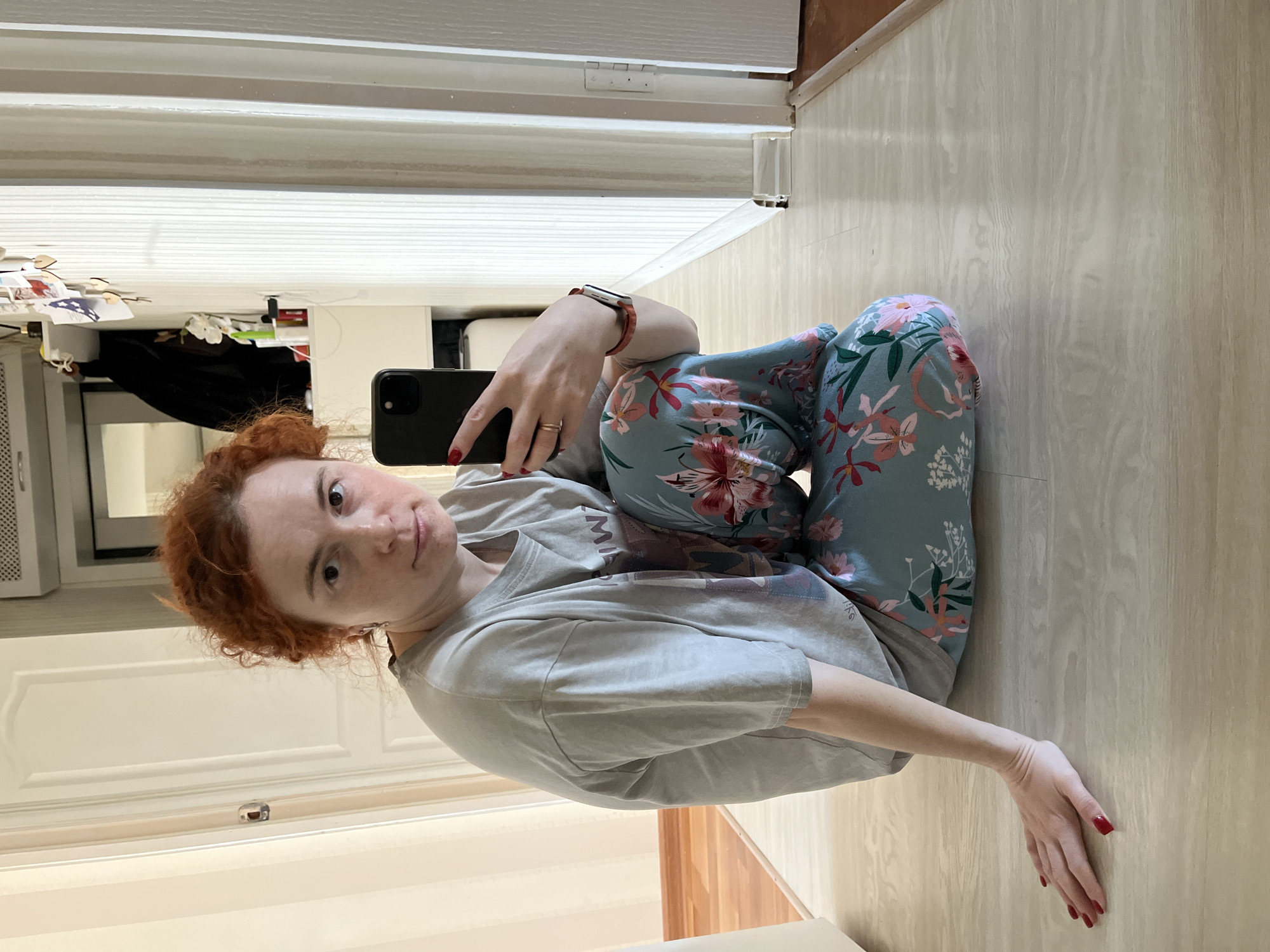 A red-haired woman in a gray t-shirt and floral pants sits cross-legged on a light-colored wooden floor, holding a black smartphone in her left hand to take a selfie. Her left arm is extended to the side, her hand resting on the floor. She has red nail polish. The background shows a white door with a handle, a white wall, and a glimpse of what appears to be a kitchen counter with items on it. The location is indoors, possibly a residential apartment, with no specific indication of Chaikovsky, Russia.