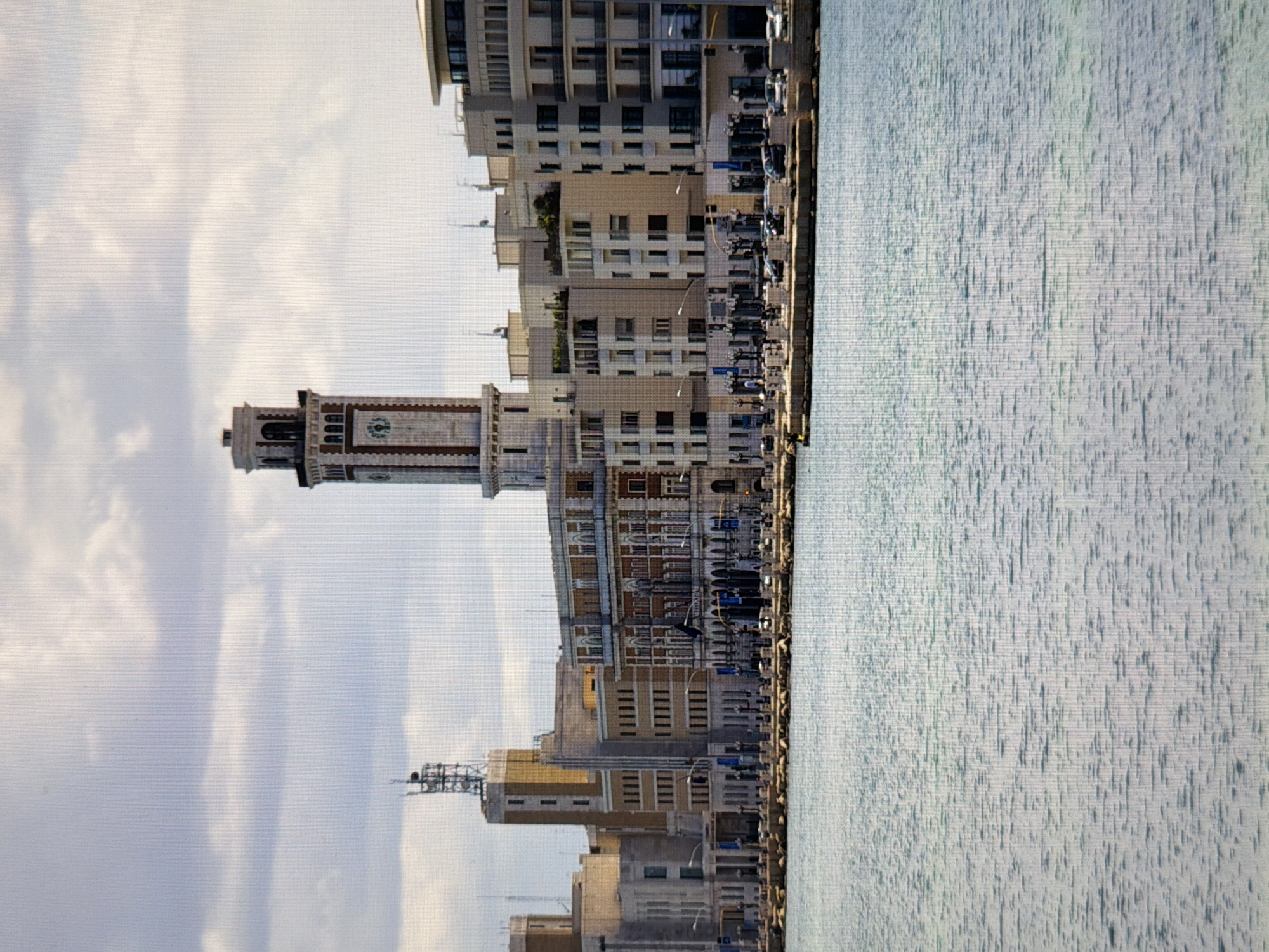 The image shows a waterfront cityscape with a prominent clock tower. The buildings are varied in style, some appearing older with ornate details and others more modern and minimalist. A body of water, likely a sea or large lake, occupies the foreground, with calm, rippling surface. A stone embankment separates the water from the buildings. The sky is partly cloudy. There are no people clearly visible in the image, though some vehicles are present along the waterfront promenade. The architecture suggests a European coastal city. The request to identify the location in Kyiv, Ukraine, cannot be fulfilled as the image does not depict any recognizable landmarks or characteristics of Kyiv.