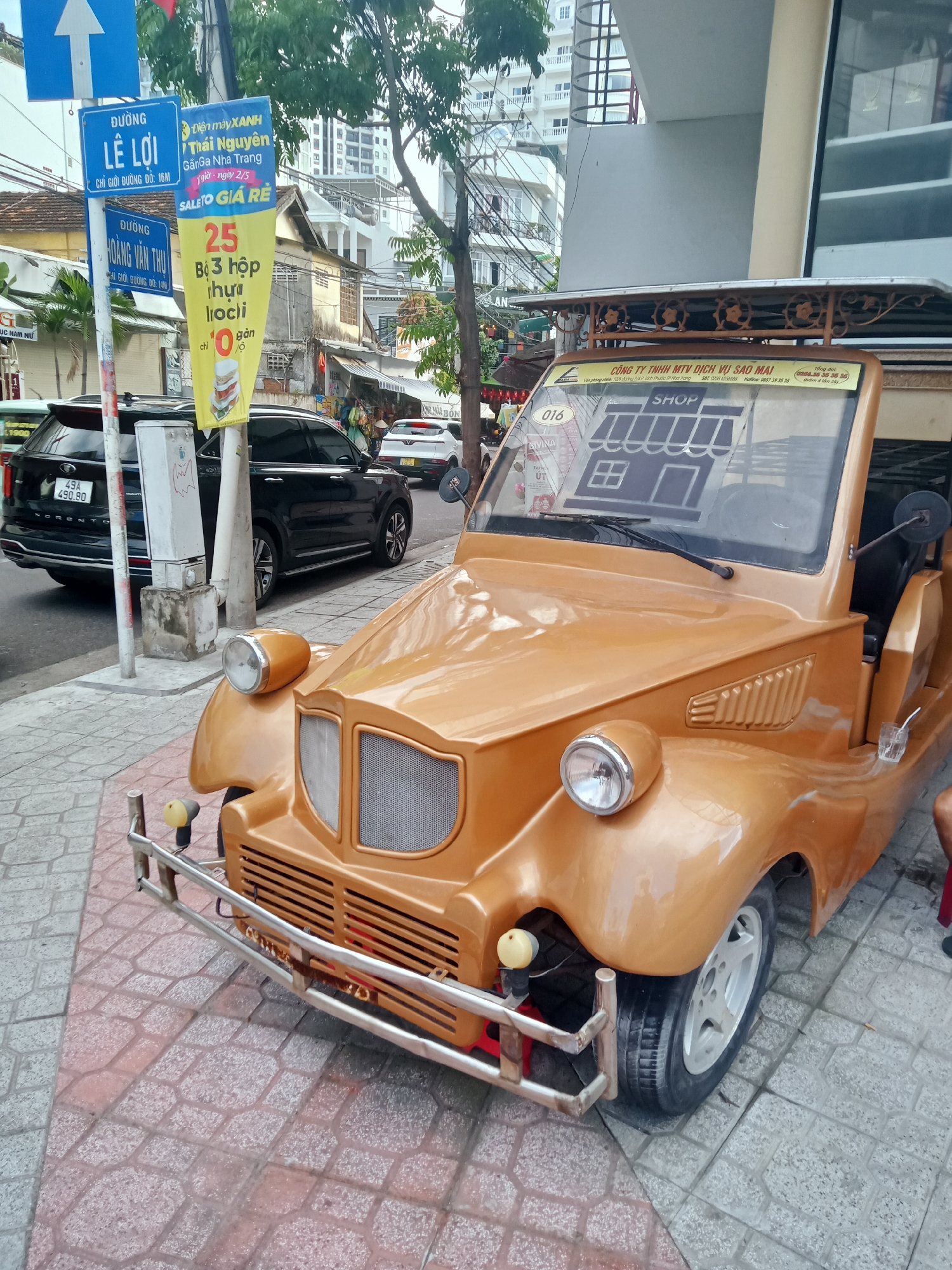The image shows a vintage-style, open-top, golden-yellow electric car parked on a patterned sidewalk in Nha Trang Ward, Vietnam. The car features chrome bumpers, large headlights, and a sticker depicting a shop on its windshield. The company name "CONG TY TNHH MTV DICH VU SAO MAI" is visible on the car. Behind the electric car, a black SUV is parked along the street. Street signs indicating "DUONG LE LOI" and "DUONG HOANG VAN THU" are visible, along with an advertisement for "SALETO GIA RE." Buildings and trees line the background, indicating an urban street scene.