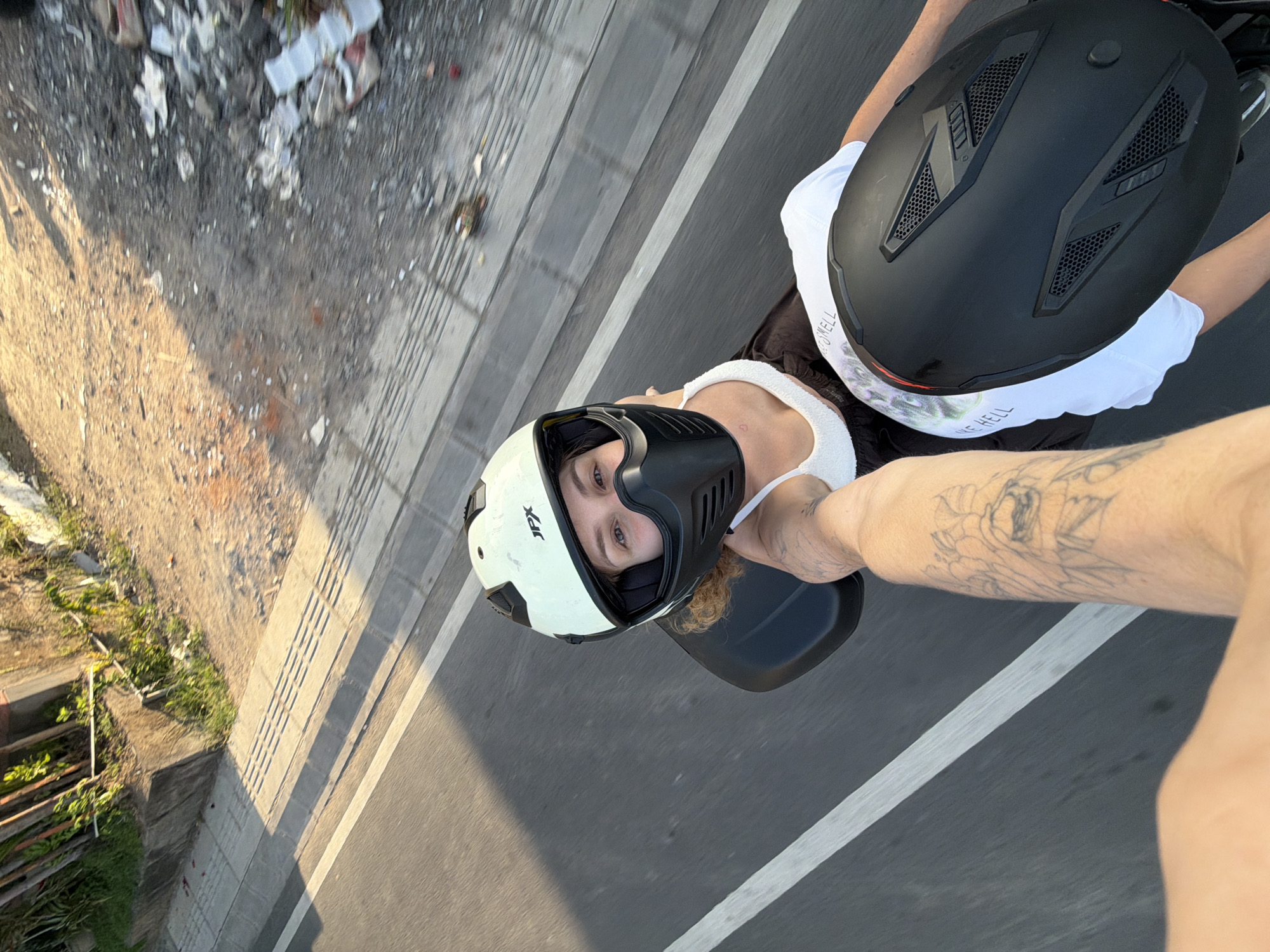 This media file captures a selfie-style shot of two individuals on a motorcycle, seemingly in motion. The perspective is from someone riding on the back, looking forward. The primary subject in the foreground is a person wearing a white tank top and a black face mask, with a white and black helmet on. Their eyes are visible, conveying a neutral expression. Their arm, adorned with a tattoo, is extended towards the camera.
To their right, another individual is seated, wearing a white t-shirt and a black helmet. Their arms are also visible, suggesting they are controlling the motorcycle. The background reveals a roadside scene with a dirt path on the left, littered with debris, and a paved road with white lines on the right. Green vegetation is visible along the edge of the dirt path.
The lighting suggests it is daytime, likely late afternoon or early morning due to the warm tones and shadows. The setting appears to be a street in Mengwi, Indonesia, given the provided location context. The overall mood is casual, possibly indicating a recreational ride or travel. No other significant text is visible besides the brand name on the helmets and potentially on the t-shirt of the person in the background.