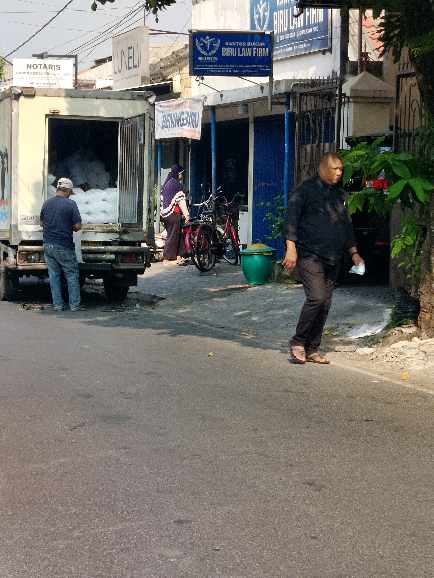 A man in a black long-sleeved shirt and dark pants walks on a sidewalk next to a street in Mojokerto, Indonesia. He is holding a white mask in his right hand and wearing sandals. To his left, a truck is parked, with its back doors open, revealing stacked white bags. A man in a blue shirt and jeans is standing by the truck, facing into it. A woman in a purple headscarf and patterned shawl walks away from the truck, also heading towards a building with blue doors. The building has signs indicating "BENING BIRU" and "BIRU LAW FIRM". Bicycles are parked in front of the building. The scene is illuminated by sunlight, casting shadows on the ground.