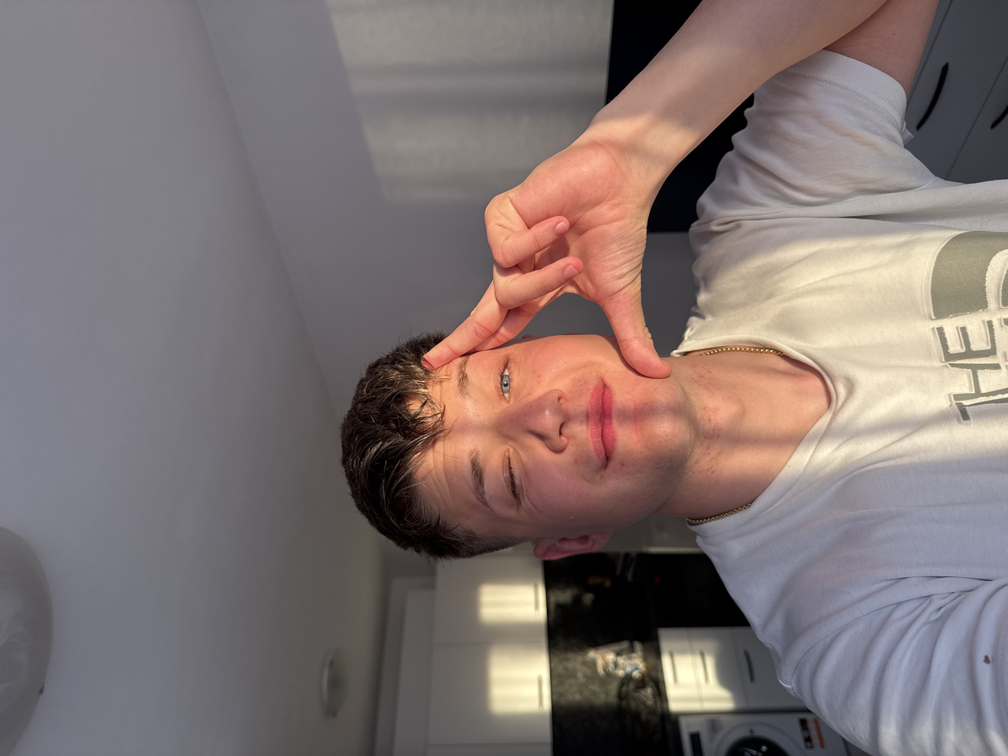 A young man with dark hair and blue eyes is posing indoors. He is wearing a white t-shirt and a gold chain. He is holding his hand up to his forehead, with his fingers making a triangle shape. The background includes white cabinets, a dark countertop, and a washing machine. The location in the city of Plovdiv, Bulgaria cannot be determined.