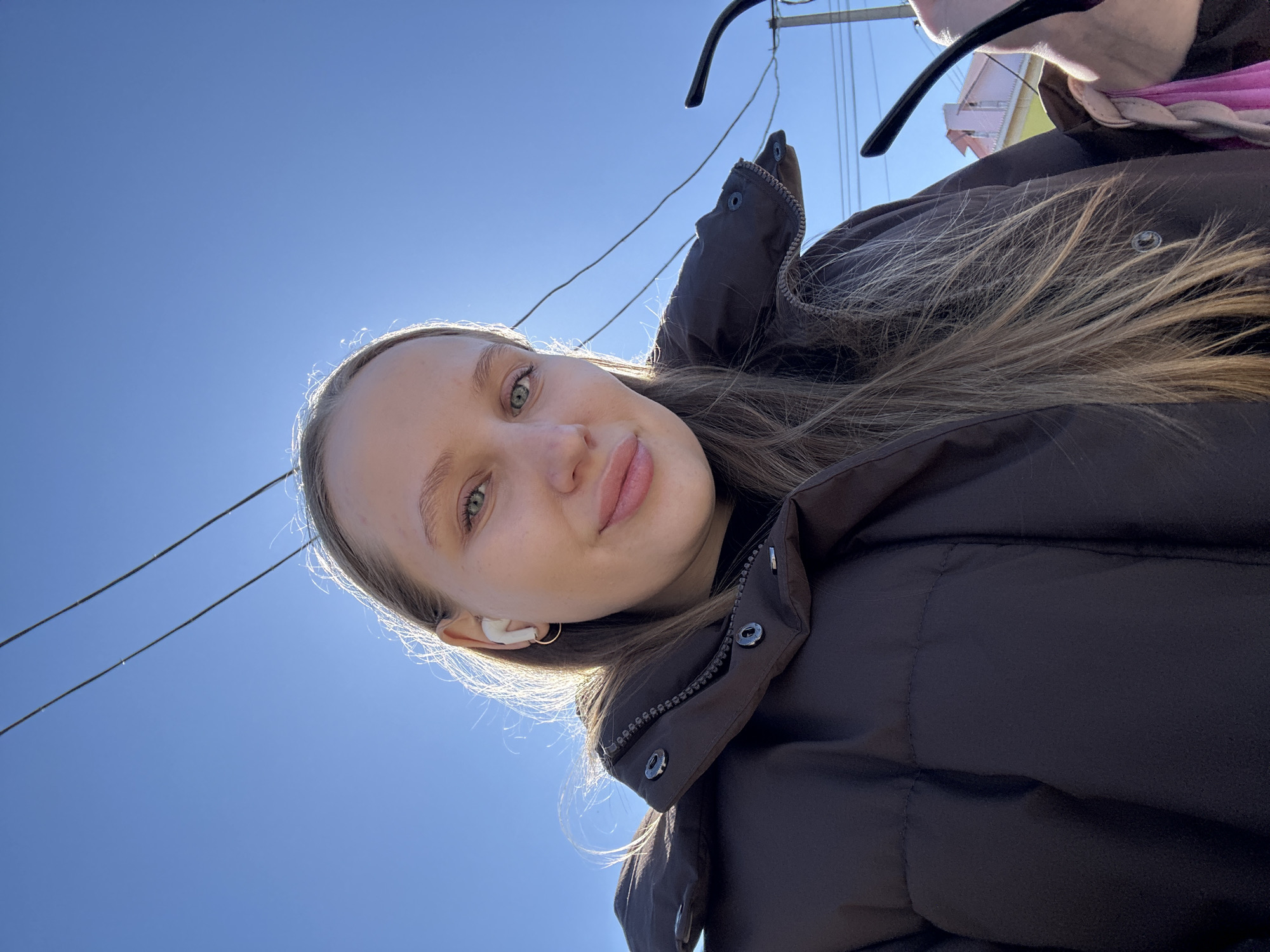 A young woman with light skin and brown hair, wearing a dark brown puffer jacket and a white wireless earbud in her left ear, takes a selfie. She is slightly smiling. Above and behind her is a clear, bright blue sky. In the background, there are power lines and the corner of a building is visible with a pink accent. Sunglasses are visible at the top of the frame. The location is likely Chaikovsky, Russia.
