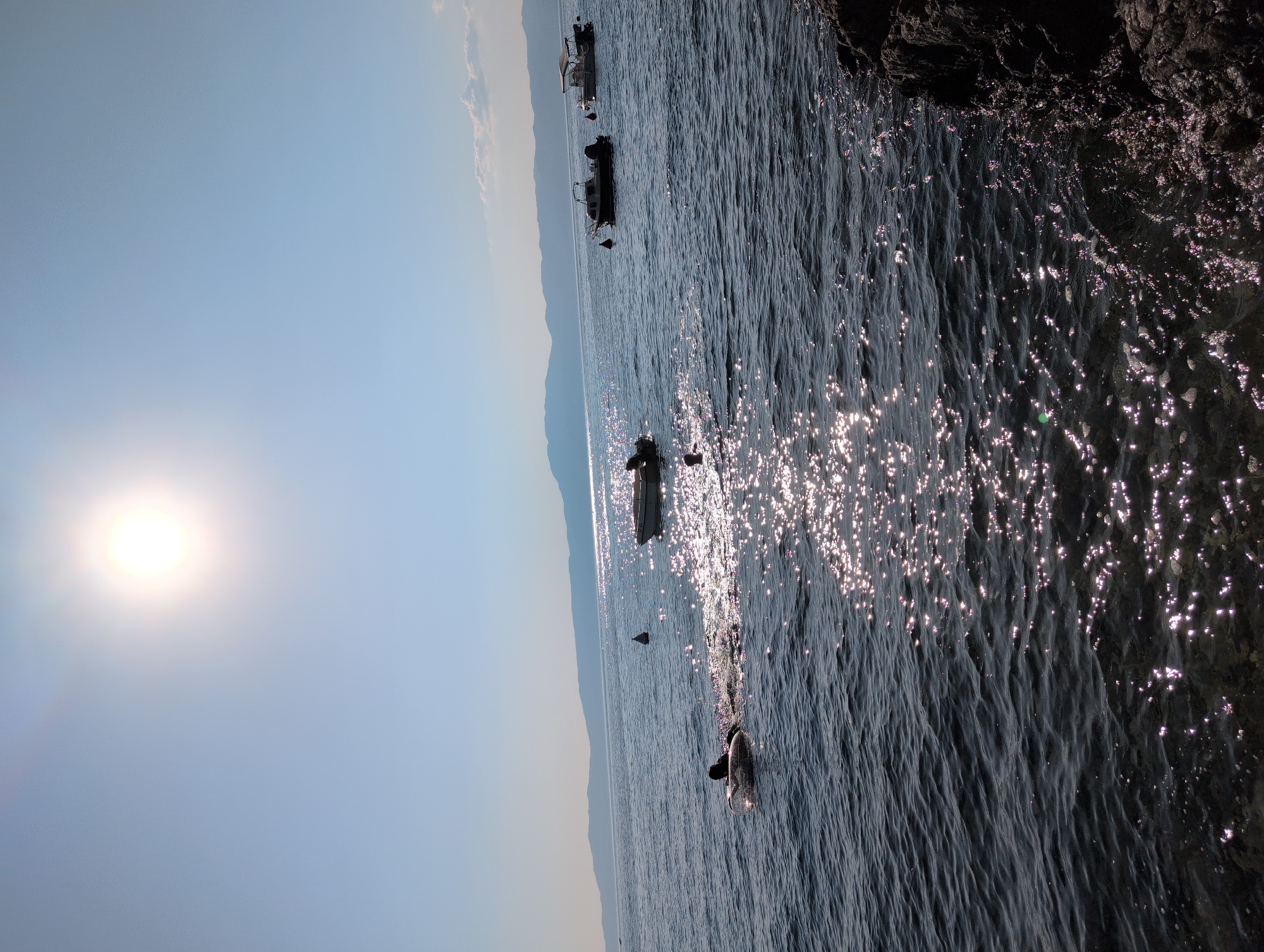 This is a sunny, outdoor, eye-level shot of the Adriatic Sea near Kraljevica, Croatia. The sun is bright and high in the sky, casting a glare on the water. Several small boats are anchored offshore. In the foreground, a person is swimming with an inflatable tube. The water is dark blue and calm, with sunlight glinting off its surface. Rocky shoreline is visible on the right side of the frame. In the background, a range of mountains is visible on the horizon. The sky is clear and blue.