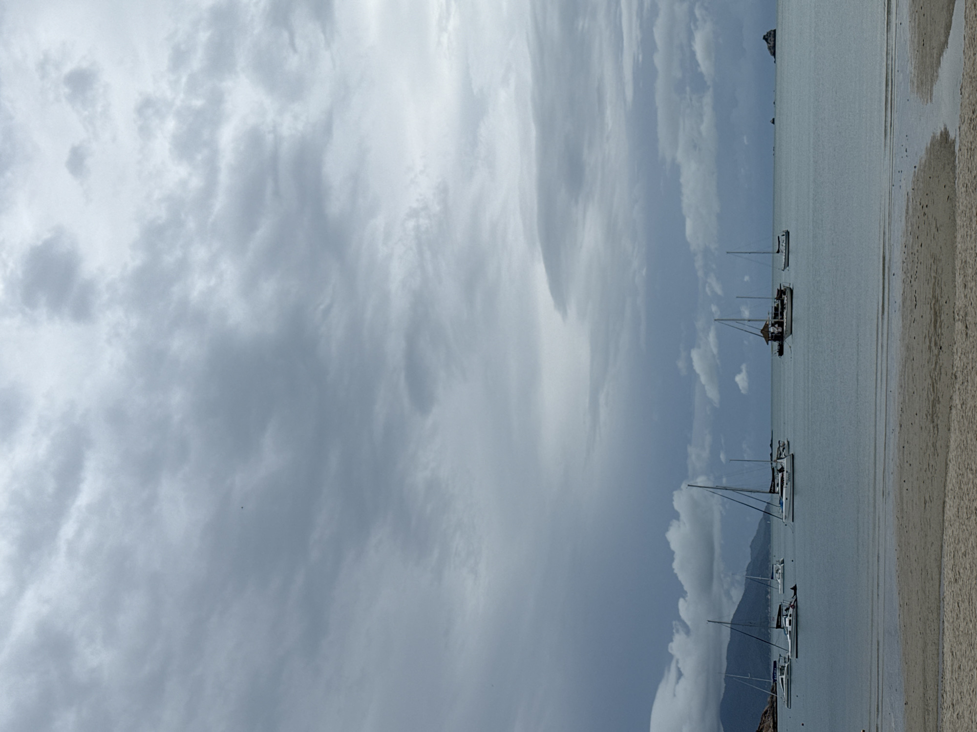 The image depicts a serene and overcast seascape, likely in the Ko Samui District of Thailand. The foreground features a sandy beach with gentle ripples of water flowing across it, suggesting a receding tide. Further out, the tranquil sea is dotted with several sailboats and a larger boat with a cabin. These vessels are anchored, indicating a peaceful anchorage. In the background, a range of lush green mountains rises majestically, their peaks partially obscured by the dramatic, cloudy sky. The overall atmosphere is calm and contemplative, with the diffused light creating a soft, muted color palette.
