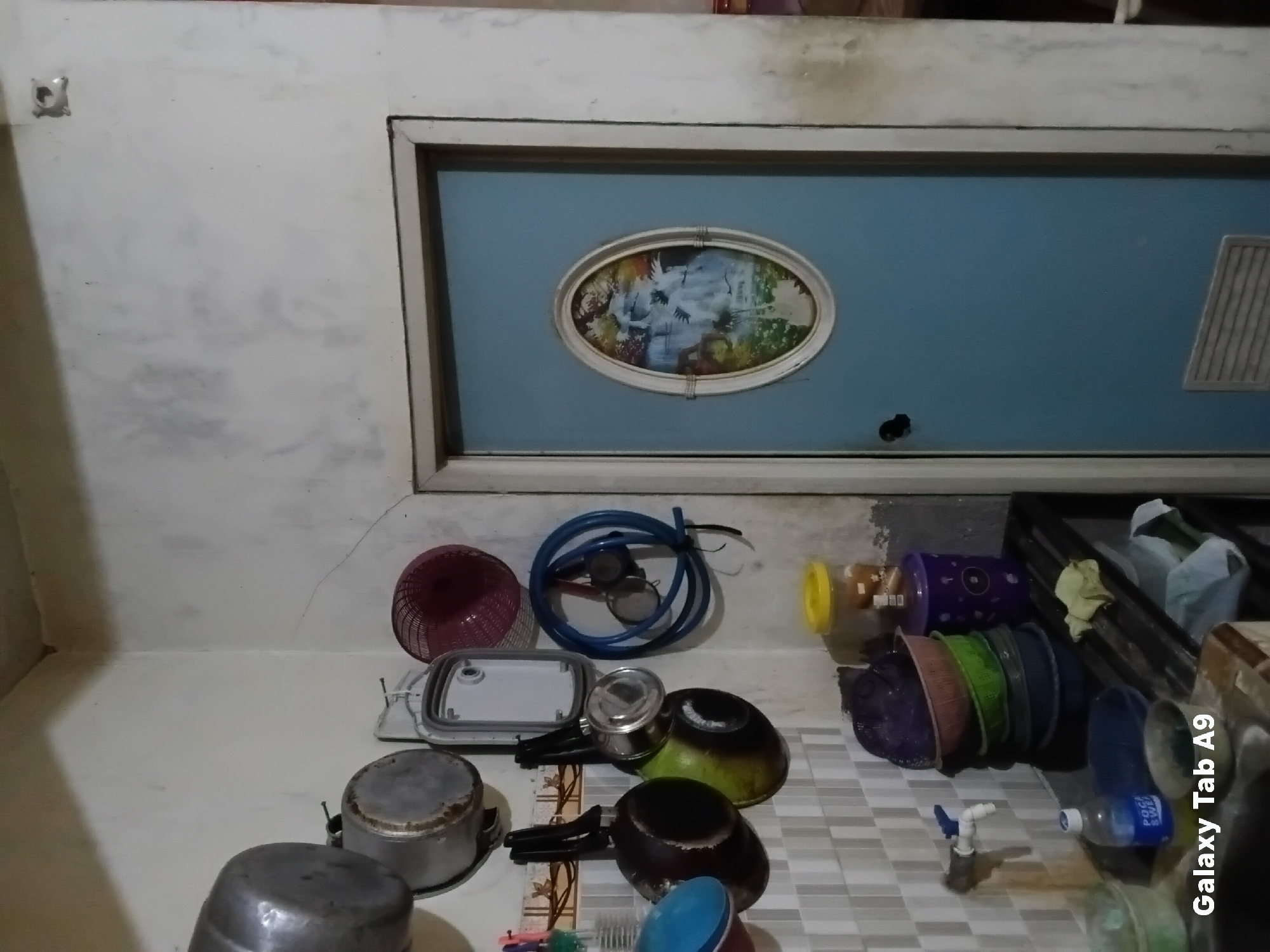 The image shows a corner of a kitchen with kitchenware hanging on the wall and stacked on a counter. On the left, several metal pots and pans are hung on hooks. Below them, on a tiled backsplash, a faucet is visible with a blue handle. A bottle of Pocari Sweat is on the counter next to a pile of stacked bowls in various colors, and a yellow-lidded container. A blue door with an oval-shaped framed picture of a waterfall is on the right side of the image. The walls are rough and textured, with some discoloration. The overall impression is of a functional, albeit somewhat cluttered, kitchen space. The text "Galaxy Tab A9" is visible in the lower-left corner of the image. The image does not contain enough information to determine the specific location within Trenggalek, Indonesia.