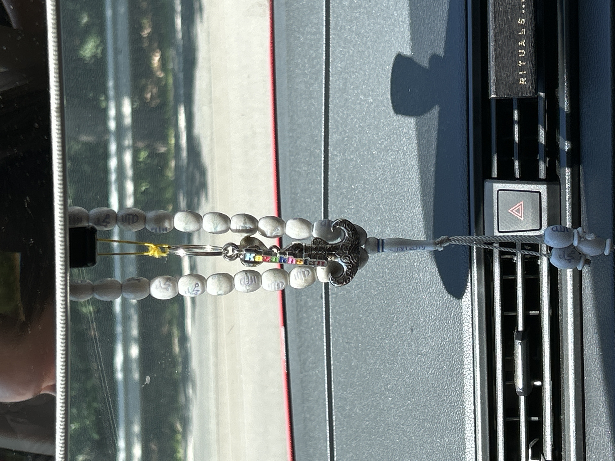 A hanging rosary adorns the rearview mirror of a car. The rosary is made of light-colored beads, some of which appear to have subtle markings. A silver-toned anchor charm, decorated with colorful segments, hangs from the rosary. Below the anchor, the rosary continues, with a white tassel-like embellishment at its end. The car's dashboard is visible in the foreground, featuring air vents and a hazard light button. A logo or text reading "RITUALS..." is present on a dark rectangular element to the right of the air vents. The scene is illuminated by natural light, casting shadows on the dashboard. The reflection in the rearview mirror shows a blurred outdoor environment, suggesting the car is likely parked or moving on a road. Given the location of Reichshof, Germany, it is probable this is a residential or commercial area within the town.
