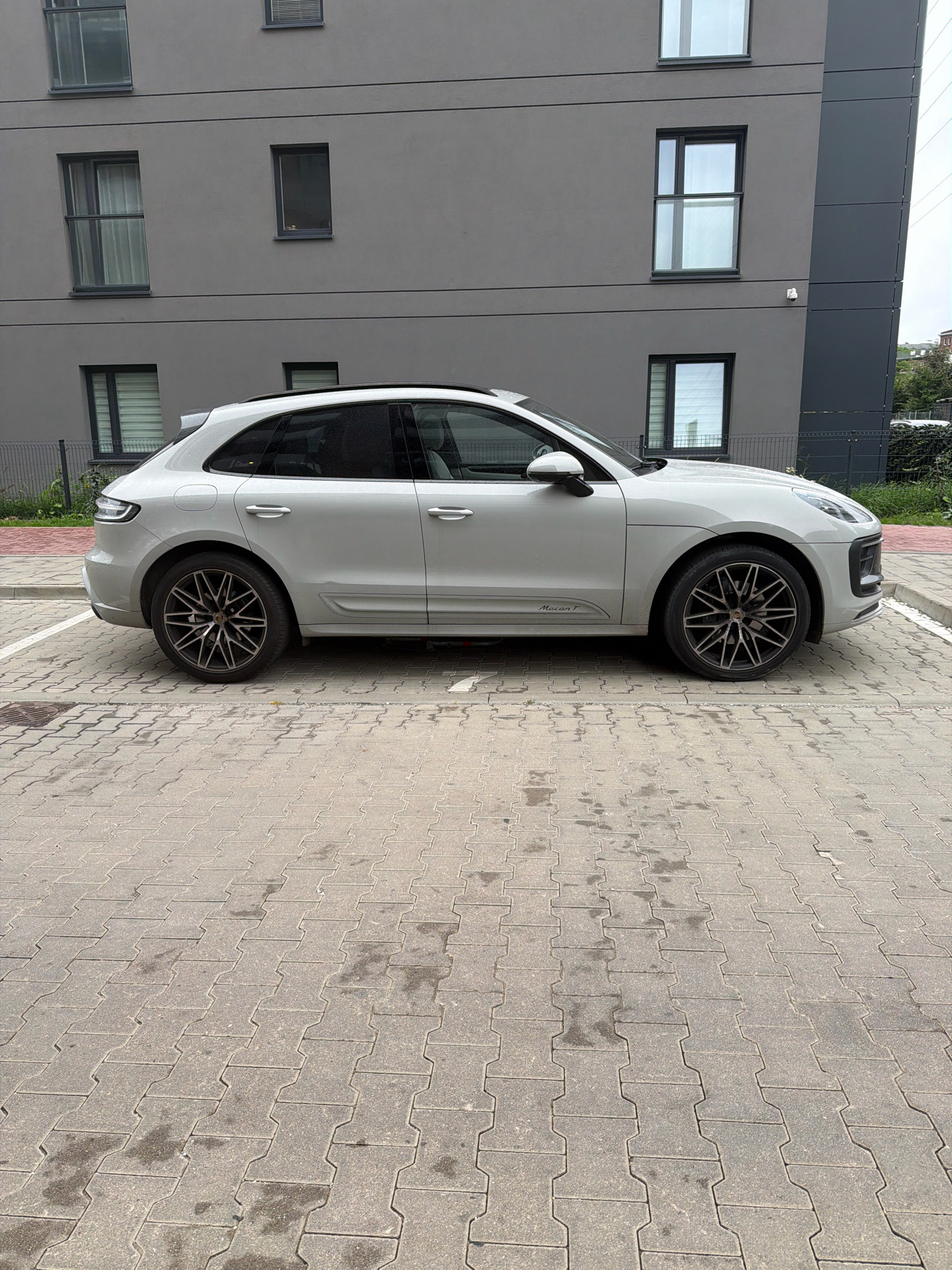 A silver Porsche Macan is parked on interlocking concrete pavers in front of a modern, gray, multi-story building. The building features several rectangular windows with dark frames. The car is viewed from its side, with its doors closed and no occupants visible. A metal fence and some greenery are visible behind the car. The pavement has some damp patches. The overall scene suggests an urban residential or commercial area.