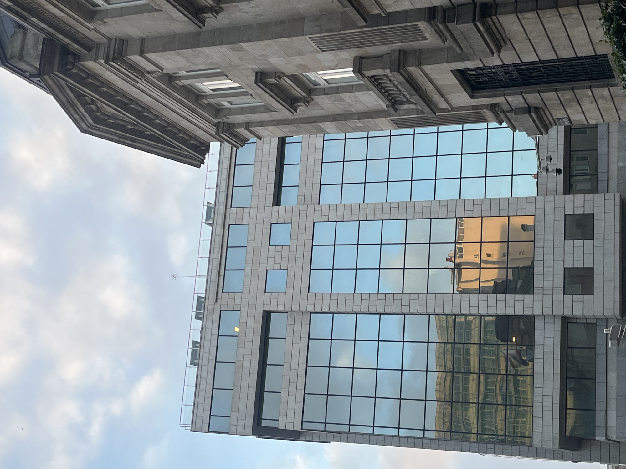 A low-angle, off-center shot shows two buildings in A Coruña, Spain, under a cloudy sky. The building to the upper left is older, made of gray stone, and features ornate architectural details like cornices and small, rectangular window openings. The building is cropped, showing only a portion of its upper floors. To the right and below is a modern building with a grid pattern of large, reflective windows and gray stone panels. The windows reflect the sky and other buildings, including a tan-colored structure. Security cameras are visible on the modern building's façade. A thin metal railing is present on the left edge of the modern building, suggesting a balcony or roof access. The image is rotated slightly to the left.