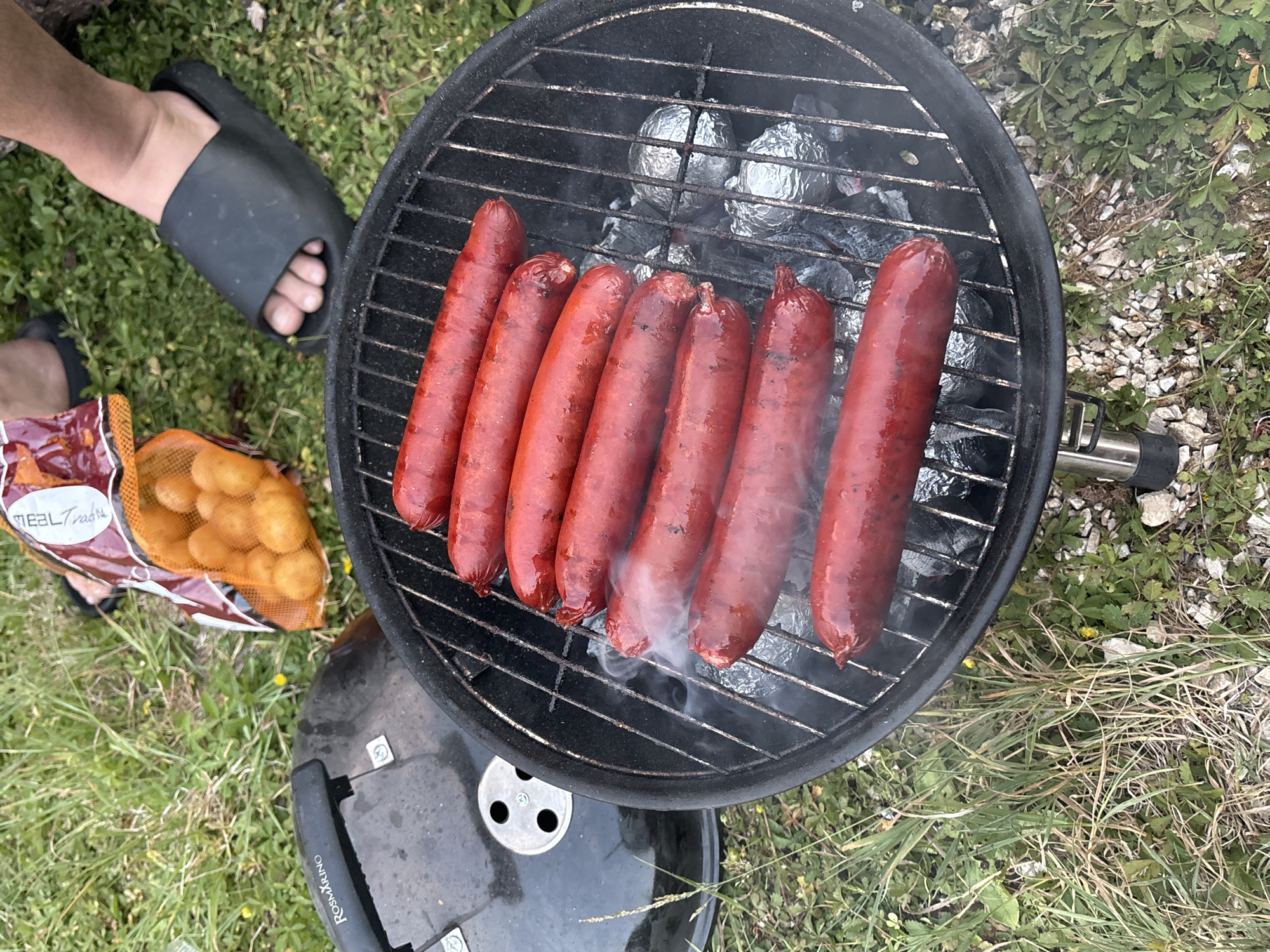 In Grahovo, Montenegro, a close-up overhead shot captures a barbecue in progress on a grassy patch. A black charcoal grill, bearing the "Rosmarino" brand on its lid, is centrally placed. On the grill rack, seven plump, red sausages are arranged, some emitting wisps of smoke. Beneath the sausages, several food items are wrapped in aluminum foil. To the upper right, a bag of small, orange potatoes is visible, resting on the grass. The scene is completed by the feet of two individuals wearing black slides, standing on the grass. The environment suggests an outdoor recreational setting, possibly a picnic or a campsite.