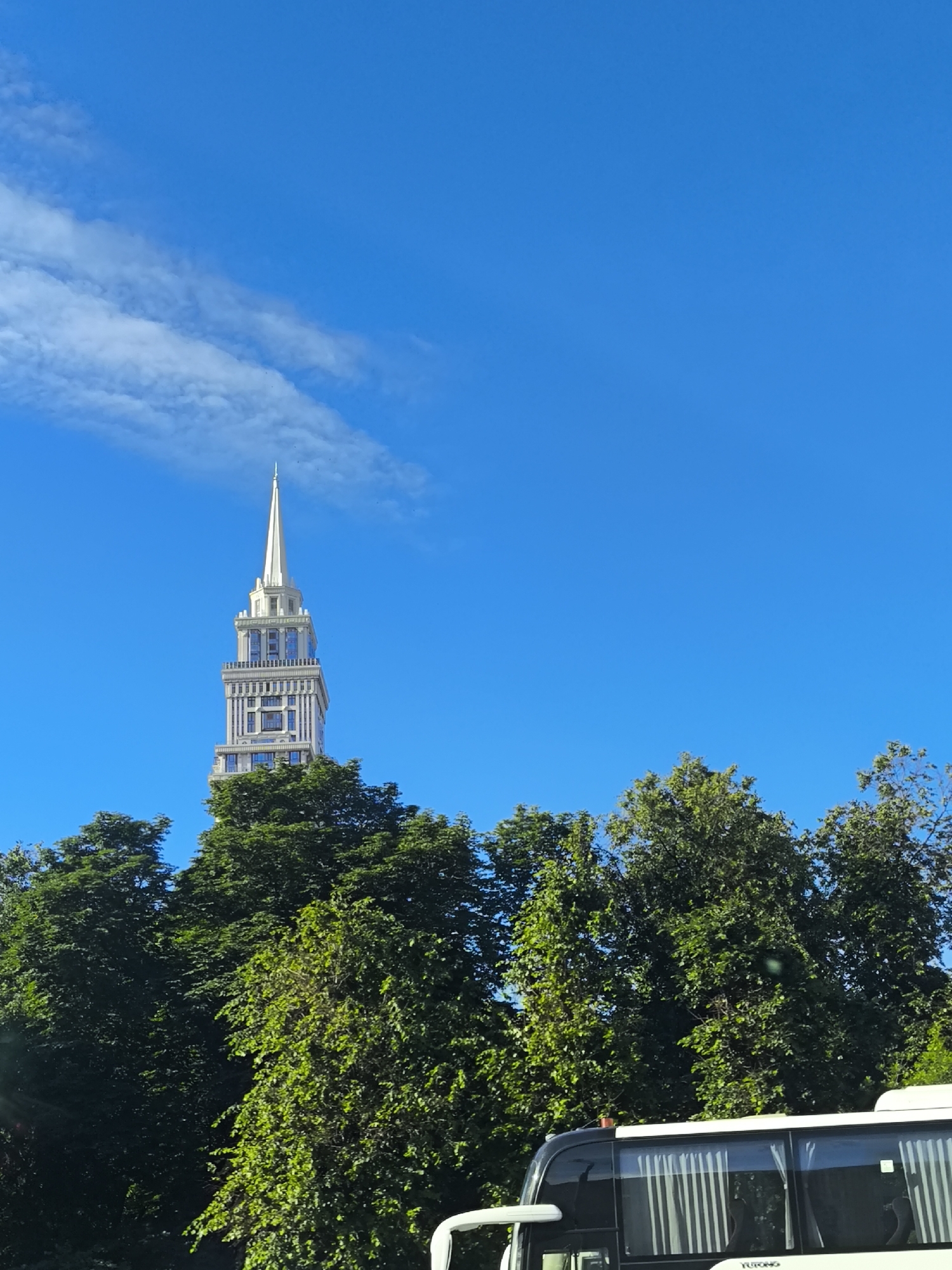 A tall, slender skyscraper with a pointed spire, in shades of white and gray with blue-tinted windows, rises above a dense line of green trees under a bright, clear blue sky with a wispy cloud. In the foreground, the top of a white bus with black trim and tinted windows is visible. The scene is set in an urban environment, likely during the daytime given the bright sunlight.