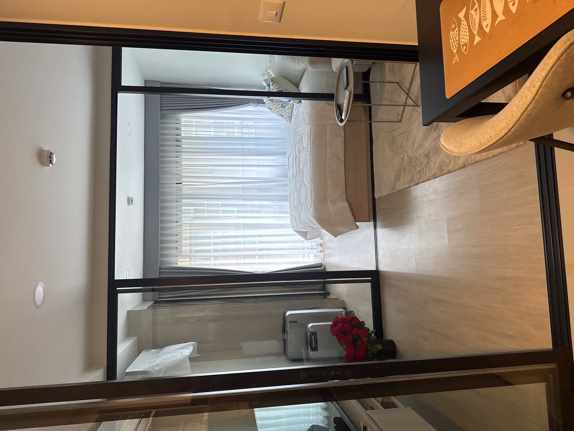 This is a photograph of a studio apartment interior, likely located in Kathu, Thailand. The room is visible through a large sliding glass door. The foreground shows a portion of a dining table with a chair. The main area of the apartment features a bed with white linens and decorative pillows, a small round side table with a remote control and a smartphone, and a sheer curtained window. To the left, two silver suitcases and a vase of red roses are placed on a light-colored wooden floor. An air conditioner unit is mounted on the wall. The overall impression is a modern and tidy living space.