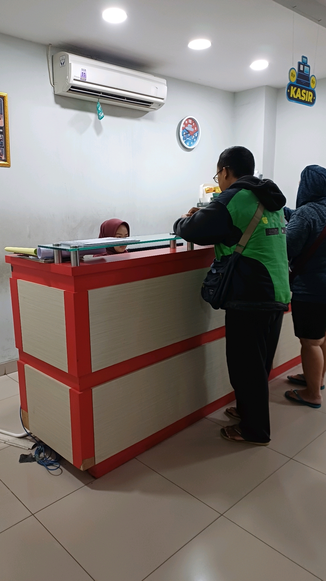 The image shows the interior of a building, likely a public service or commercial establishment, in Cibinong, Indonesia. A red and beige reception desk is prominent in the foreground, with a glass partition on top. Behind the desk, a woman wearing a maroon hijab is visible, appearing to be an employee. Two men are standing in front of the desk, interacting with the employee. One man, wearing a green vest over a black hoodie and black pants, is facing the desk and holding something. The other man, wearing a dark hooded jacket, is partially visible to the right, also facing the desk. On the wall behind the desk, there is a white air conditioner unit and a round clock with a blue and red face. A sign with the word "KASIR" (Indonesian for "Cashier") is hanging from the ceiling, indicating the function of this area. The floor is tiled in a light color. The overall scene suggests a transaction or service interaction at a cashier's station.