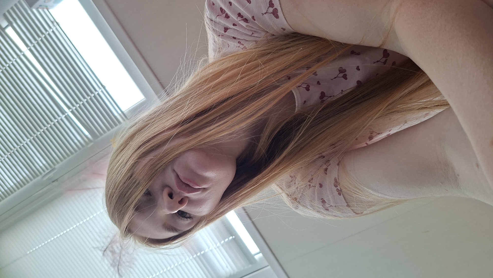 The image presents a close-up, low-angle portrait of a young individual, likely female, with long, light reddish-brown or strawberry blonde hair cascading around her face and shoulders. The subject is wearing a light-colored garment, possibly a pajama top or casual shirt, patterned with small, dark red heart shapes and subtle lines.
The visible portion of the face, from the nose down, shows a soft, neutral expression with slightly parted lips. Parts of the eyes are visible, looking upwards towards the camera. The subject's arm and shoulder are also discernible, blended into the light background.
In the background, a brightly lit window with horizontal blinds or slats is prominent, indicating an indoor setting bathed in daylight. The overall composition is rotated, suggesting the photograph was taken from below, possibly while the person was reclining or looking up. There is no specific activity depicted beyond posing for the photograph. No visible text is present. The provided location context is Chaikovsky, Russia.