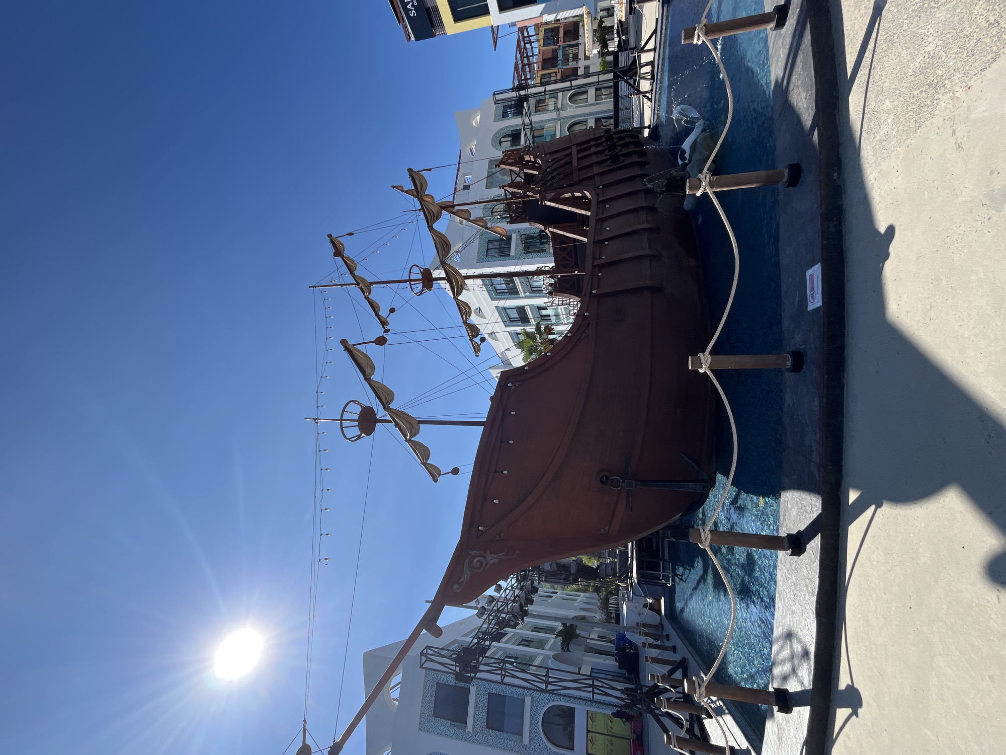 The image displays a decorative replica of a wooden pirate ship situated in a shallow pool of blue water. The ship is brown and has multiple masts with furled sails, decorative lighting strings, and railings. The pool is surrounded by a concrete walkway with rope and wooden post barriers. In the background, there are modern multi-story buildings with white and light-colored facades, featuring balconies and windows. The sky is clear and bright blue, with a prominent sun casting lens flare. There are no people visible in the scene. The location is identified as Bac Nha Trang Ward, Vietnam.