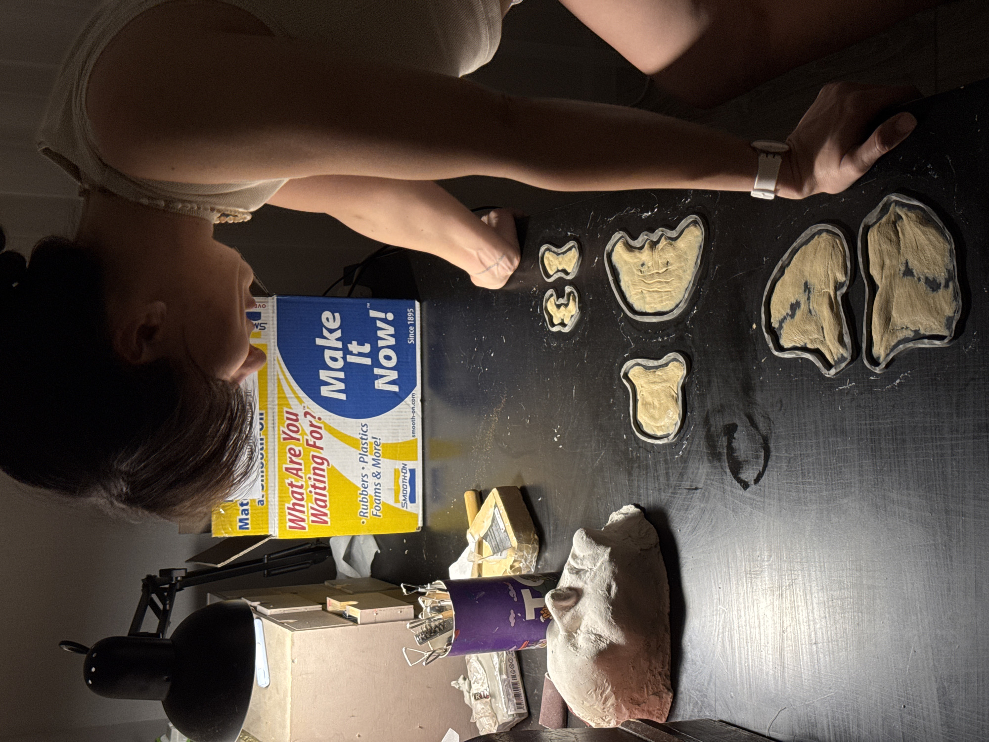 A person is positioned above a black table with several objects on it. The person is wearing a light-colored top and a white wristband. On the table, there is a box labeled "Make It Now!" featuring the text "What Are You Waiting For?" The table also contains several molds, including molds of face parts, and a three-dimensional model of a face. There are also various tools in a purple cup. A black desk lamp is visible in the lower left of the image. The environment is dimly lit. The location is not mentioned in the image.