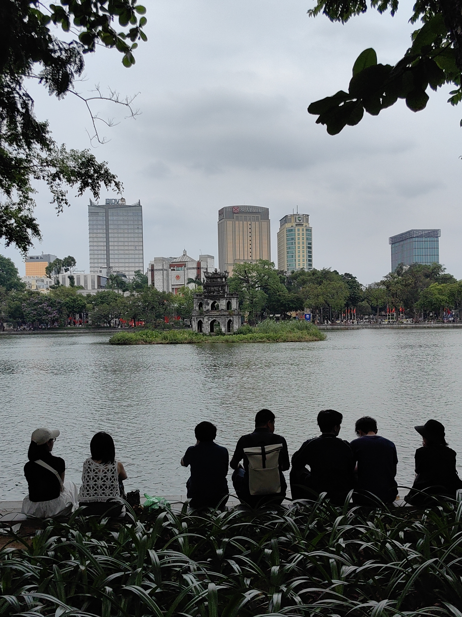 The image captures a panoramic view of Hoan Kiem Lake in Hà Nội, Vietnam, under an overcast sky. In the immediate foreground, dark green leafy plants are visible, partially obscuring a low wall or embankment. Six individuals are seated on this wall, seen from behind, gazing out over the calm waters of the lake. One person on the left wears a white cap and white skirt, while another has a distinctive white crocheted top. One person in the middle has a white backpack.
In the mid-ground, a small, tree-covered island features the historic Turtle Tower, an ancient, multi-tiered structure with arched openings. The lake's surface appears tranquil, reflecting the muted light of the sky.
The background reveals a modern urban skyline surrounding the lake. Several high-rise buildings are visible, including one on the left prominently displaying "BIDV" at its top, and another slightly lower building with "Fairmont" visible. Further to the right, other tall buildings feature bank logos, including "SeABank" and another with a red, indistinct logo. The far bank of the lake is lined with trees and what appear to be red flags, suggesting a public area or promenade. Overhanging tree branches frame the top and upper left of the photograph. The scene blends natural beauty and historical architecture with contemporary city development, under soft, diffused lighting typical of a cloudy day.