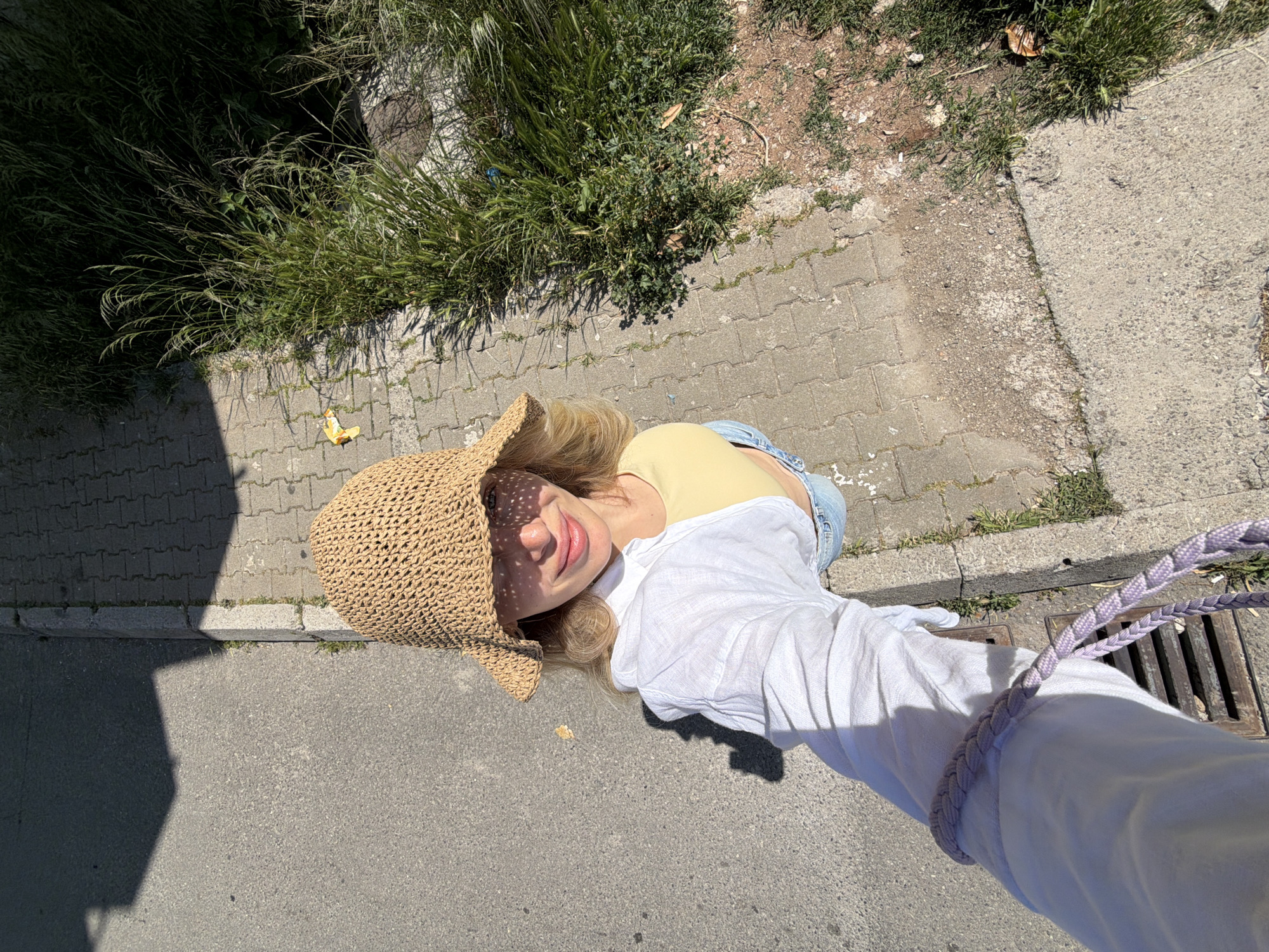 This high-angle selfie captures a young woman enjoying a sunny day outdoors in Budva, Montenegro. The woman, with blonde hair, is smiling up at the camera. She is wearing a woven straw sun hat, which casts a pattern of sunlight and shadow across her face. Her attire includes a light yellow top, light blue jeans, and a white, long-sleeved button-up shirt or jacket worn over her top.
The setting combines urban elements with natural growth. The woman is situated on a paved surface consisting of grey rectangular pavers, bordered by patches of tall, green grass and weeds. To her right and in the foreground, the ground transitions to dark asphalt. Strong, defined shadows cast by objects and the woman herself indicate bright, direct sunlight, suggesting it is midday or early afternoon.
Notable details include a light purple, braided strap or handle, likely from a bag, visible in the bottom right corner, extending towards a metal drain grate. A small, yellow/orange piece of litter lies on the paved ground near the grass. The vegetation appears somewhat wild, contrasting with the man-made pavement. The woman's expression conveys a sense of happiness and ease, fitting for a warm, sunny day. No visible text is present in the image.