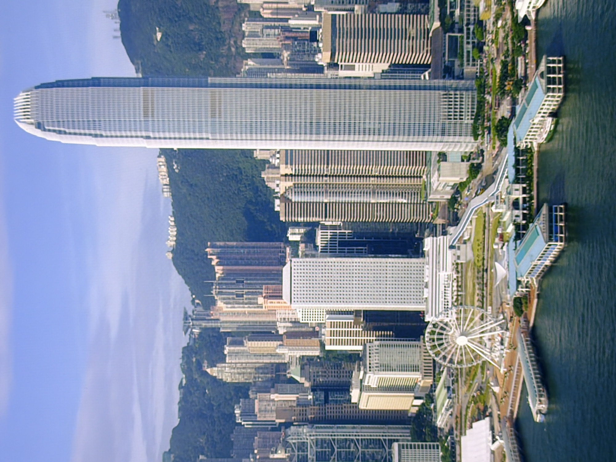 The image depicts a panoramic view of a modern cityscape with numerous skyscrapers. A prominent, tall, glass-facade skyscraper with a distinctive curved top dominates the center-right of the frame, likely being the most notable structure. To its left, a cluster of other high-rise buildings of varying architectural styles and heights forms a dense urban landscape. In the foreground, a body of water is visible, with piers and walkways extending into it. A large Ferris wheel is situated on the waterfront, suggesting a recreational area or attraction. Lush green hills or mountains form the backdrop to the city, under a clear blue sky with some scattered clouds. The scene appears to be a bustling metropolis, characterized by its impressive architecture and waterfront development. The image is a static, aerial shot, capturing the city from a high vantage point. There are no people or animals visible in the image. This scene is not located in Kyiv, Ukraine, but rather it is a depiction of Hong Kong.