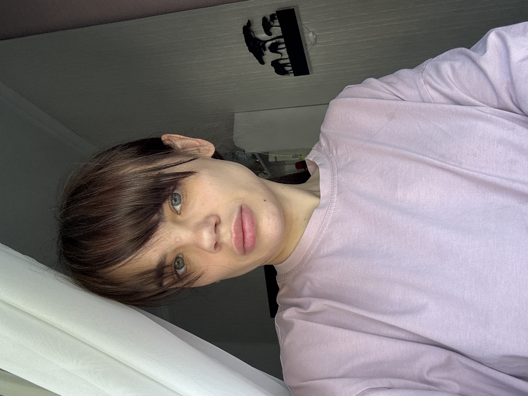 A woman with brown hair and fair skin is depicted in a medium close-up shot. She is wearing a light purple t-shirt. Her gaze is directed toward the viewer. The background is indistinct, showing a ceiling with a light fixture that appears to be a black silhouette of elephants. Part of a white curtain is visible on the left. The location and city cannot be determined from the provided information.
