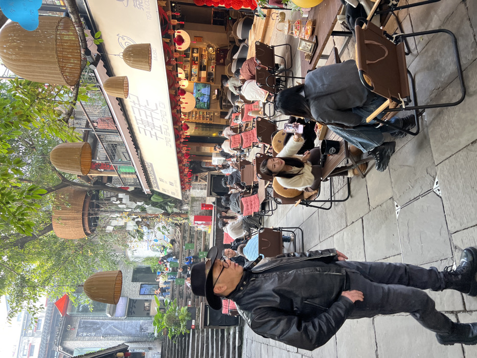 An outdoor scene in a pedestrian area of Yuzhong District, China, depicts a commercial courtyard setting. The ground is paved with rectangular stone slabs. The environment is partially shaded by lush green trees, from which multiple large, brown, wicker-basket-shaped decorative elements are suspended. One particularly large wicker structure appears to be a treehouse-like seating area or permanent decoration with wind chimes hanging beneath it.
In the foreground, a man of East Asian descent, appearing middle-aged to elderly, walks from right to left. He wears a black cowboy hat with a metallic detail, a black leather motorcycle jacket, dark trousers, and black lace-up boots. He has glasses and his left hand is in his pocket. He is looking slightly to his left.
To the man's right (viewer's right), an outdoor cafe, identified by the Chinese characters "ZAO SHU ZAO QI" (早树早起) and "TEA & COFFEE MUSIC" on its awning, occupies the midground. The shopfront is adorned with numerous red and white lanterns. This cafe features an array of brown folding chairs with red cushions and small wooden tables, arranged for outdoor seating. Approximately 15-20 individuals are seated at these tables, engaged in various social activities.
Two women are prominent in the midground, seated at a table on the right. One woman, facing the camera, wears a light-colored fluffy top and dark pants. She holds a smartphone extended to capture a selfie or photo. She has long dark hair and is smiling. The other woman, with her back mostly to the camera, wears a grey sweater and distressed blue jeans. A dark mug and a clear container with snacks are on their table. Inside the cafe, a television screen is visible, displaying content.
To the man's left (viewer's left), a wide stone staircase ascends to an upper level, alongside which tiered wooden or stone benches create an amphitheater-like seating area. Several individuals are seated on these benches. Buildings with multi-story, traditional Chinese architectural styles, featuring tiled roofs and balconies, line the background on both sides of the courtyard. The overall atmosphere suggests a lively commercial or recreational hub.