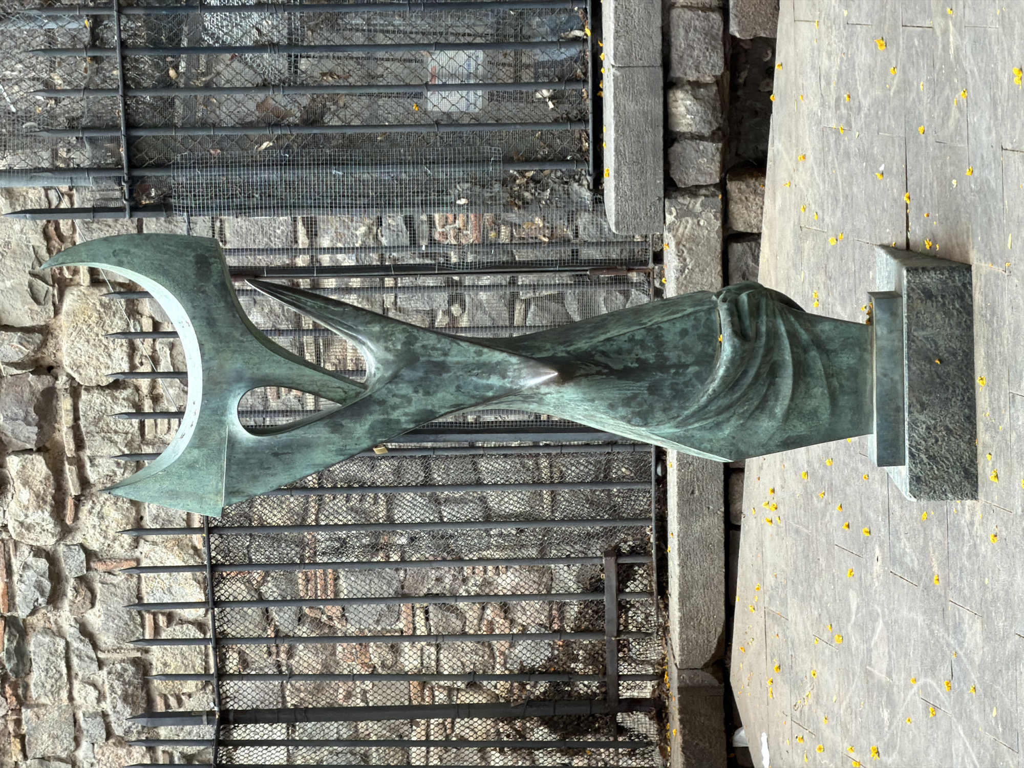 The image displays an outdoor scene in what appears to be Barcelona, Spain, featuring a striking abstract sculpture. The central subject is a tall, slender sculpture made of patinated bronze or a similar metal, exhibiting a distinct greenish-blue hue. Its form is dynamic and abstract, characterized by flowing, curved lines and sharp points, resembling a stylized bird or a sweeping wave. The sculpture rests on a dark, rectangular base.
In the background, to the left of the sculpture, is a sturdy stone wall composed of irregularly shaped, light-colored blocks. A dark metal grille or fence spans horizontally behind the sculpture, positioned in front of the stone wall. This fence features several horizontal bars, some adorned with upward-pointing spikes, and a finer mesh behind them. Beyond the fence, some natural ground with debris or sparse vegetation is visible.
The ground in the foreground and to the right of the sculpture is paved with large, light-grey stone slabs. Scattered across these paved surfaces and on top of the metal fence are numerous small, bright yellow leaf-like debris or flower petals, adding a vibrant detail to the otherwise muted tones. The scene is brightly lit by clear daylight, suggesting it is daytime. There is no visible text or human activity in the image.
