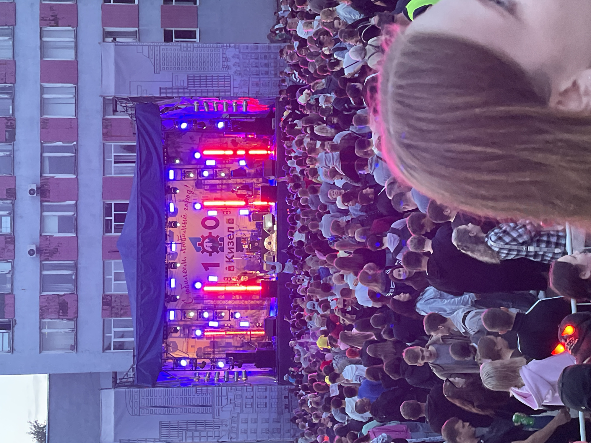 In an outdoor urban setting, likely an event or festival, a large crowd of people is gathered in front of a stage erected in front of a multi-story building. The building appears to be made of concrete and features rows of windows with some showing signs of wear. The stage is covered with a dark canopy and illuminated by vibrant red and blue stage lights. A banner or backdrop behind the stage displays a large "100" graphic and text in Cyrillic, suggesting a centennial celebration for the city of Kizel. Performers are visible on the stage, including a person with a microphone. The crowd consists of individuals of various ages, dressed in casual attire, many looking towards the stage. The scene is captured during daylight or dusk, with the lighting from the stage creating a dynamic atmosphere. The foreground shows the backs of heads and shoulders of people in the crowd, one individual in the extreme foreground has bright pink lighting on their hair and a portion of their face visible.