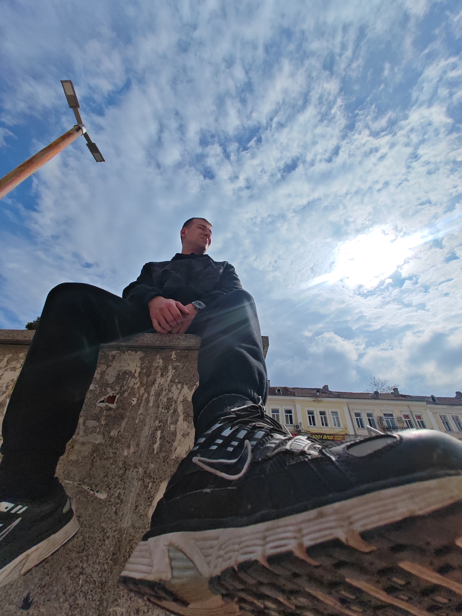 A young adult male is seated on a textured, worn concrete ledge in a commercial or pedestrian area of central Shumen, Bulgaria, captured from a low-angle perspective looking upwards. The male, with short hair, wears a black hooded top, black trousers, and black athletic shoes with white soles and accents. A silver-colored watch is visible on his left wrist. His hands are clasped in front of him, and he gazes towards his upper right, away from the camera.
The concrete ledge displays remnants of torn paper or posters on its vertical face. To the far left, a rusty orange street lamp post with a T-shaped arm holding two rectangular LED light fixtures extends into the sky.
The upper portion of the scene features a bright blue sky with scattered white, patchy clouds. The sun is visible in the upper right, casting strong light and lens flare into the frame. Below the sky, in the background, a multi-story building complex with a prominent yellow facade, white-framed windows, and reddish-brown tiled roofs is visible. A commercial sign on this building reads "1001 ВИДА" and "ЕВТИНИ СТОКИ" (1001 Kinds, Cheap Goods). Air conditioning units are attached to the building's exterior.