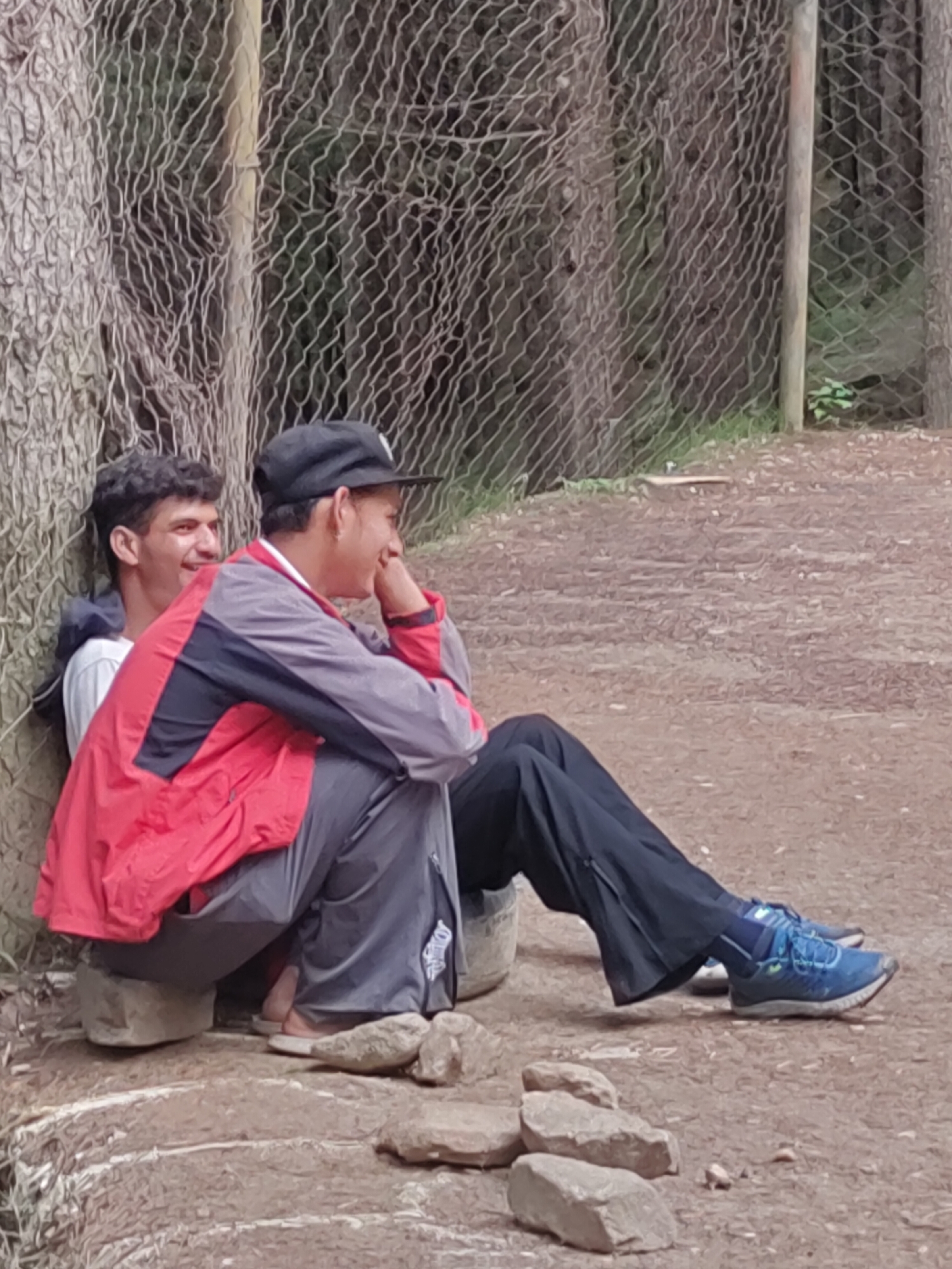 Two young men are sitting outdoors, leaning against a tree and a chain-link fence. The man on the left, wearing a white t-shirt, has his arm around the man on the right. The man on the right is wearing a red and gray jacket, dark pants, and a black baseball cap. He is resting his chin on his hand and smiling. The ground is dirt and covered with small rocks. The background shows trees and foliage. This scene is likely located in the city of Manali, India, a popular tourist destination known for its natural beauty and outdoor activities.