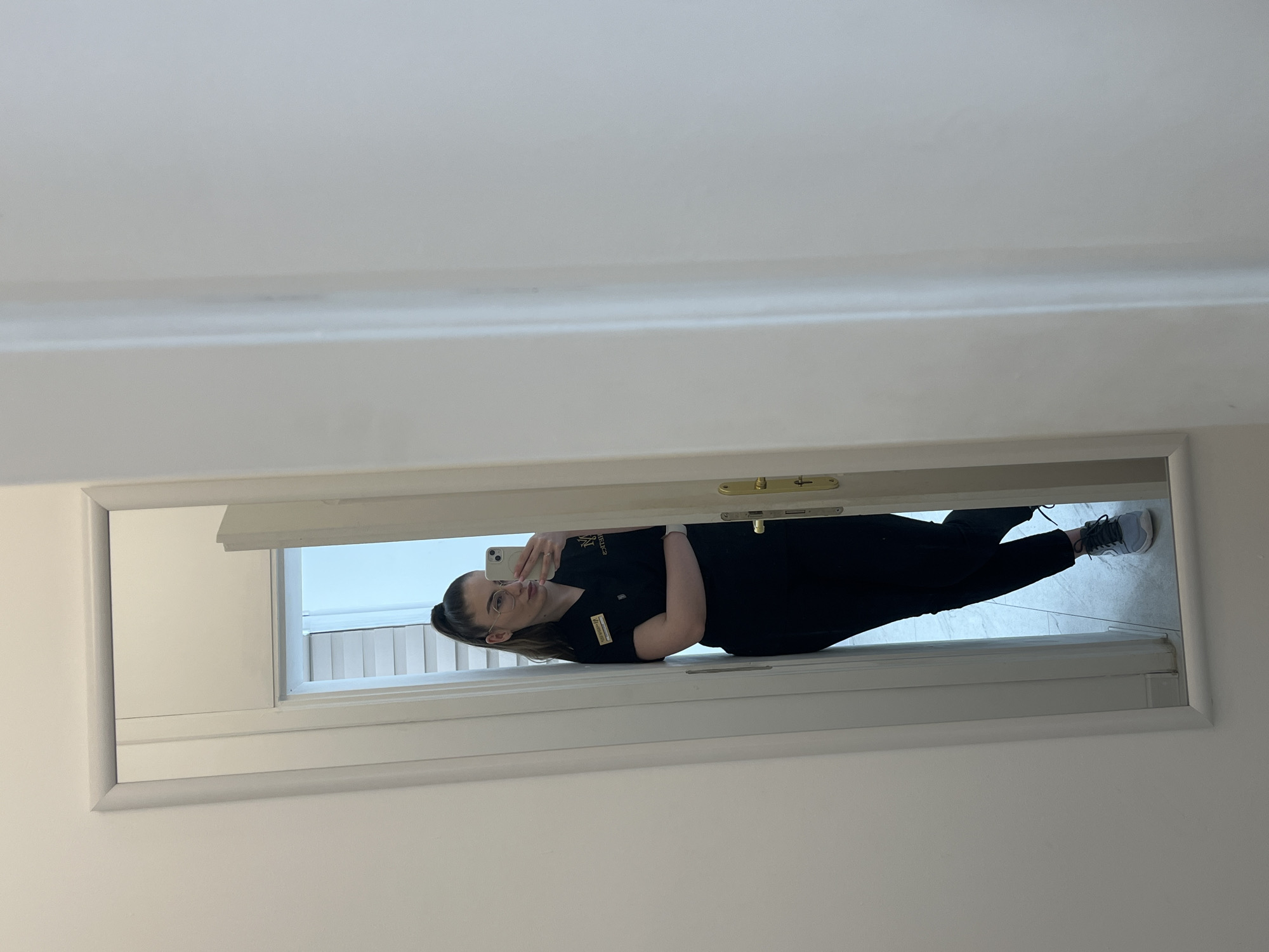 A reflection of a woman in a black uniform, with black pants and grey running shoes. She is holding a phone taking a picture of herself in a rectangular mirror. The background inside the reflection appears to have light gray tiles. The location appears to be inside of a building, Unknown, Unknown.