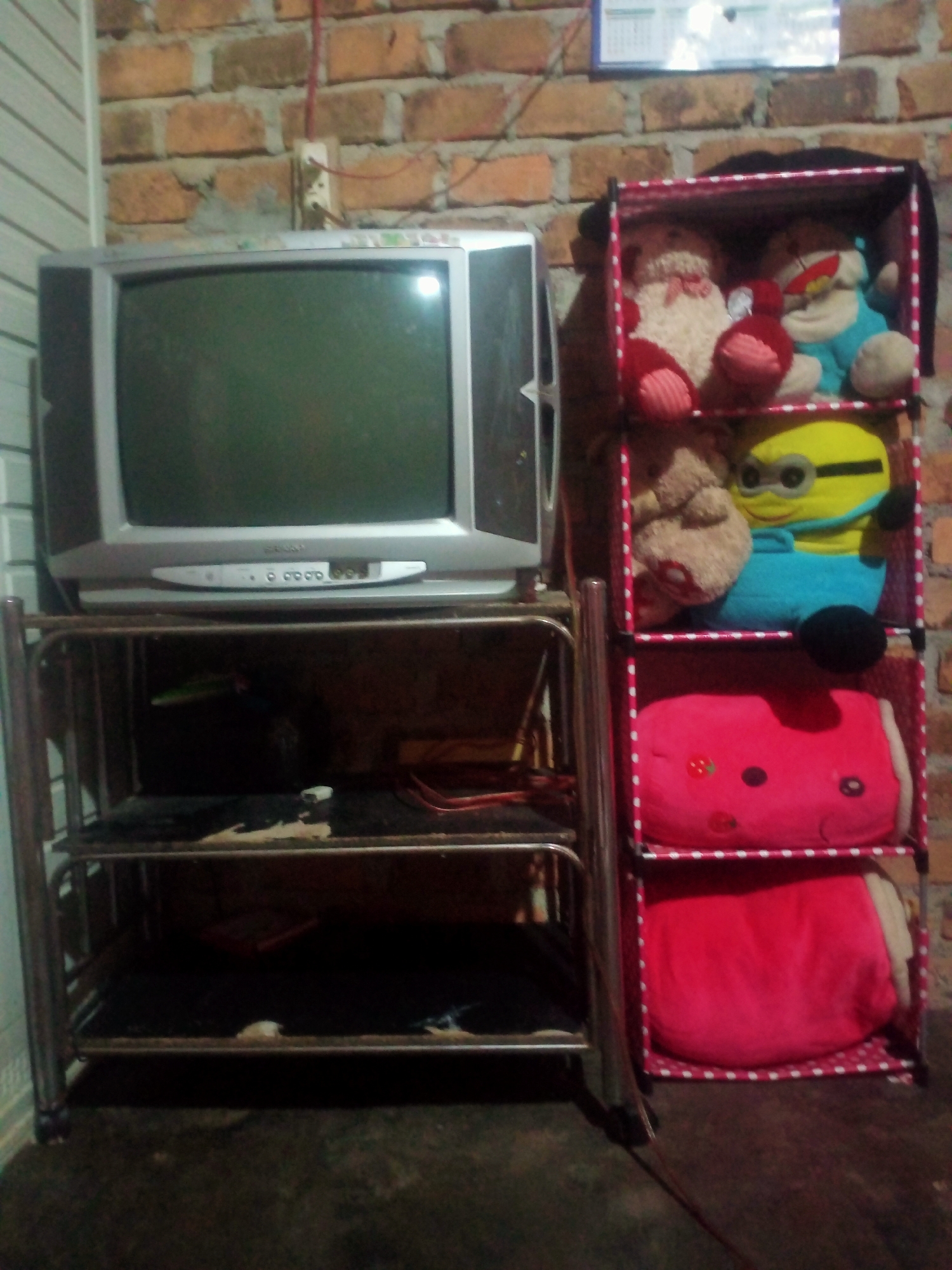 A silver CRT television set rests on a metal shelf unit. To the right of the television is a pink fabric shelving unit filled with plush toys. The top shelf of the pink unit holds two toys: one is a tan-colored teddy bear with red accents, and the other is a blue character with yellow accents and goggles, resembling a minion. The middle shelf contains a brown teddy bear. The bottom shelf is occupied by a large pink heart-shaped pillow. The television is placed on the top shelf of the metal unit, which has two additional shelves below it, both filled with dark items, likely clothing or fabric. The background consists of a brick wall and a section of vertical white siding. A red wire runs from an electrical outlet on the brick wall. The scene appears to be indoors.