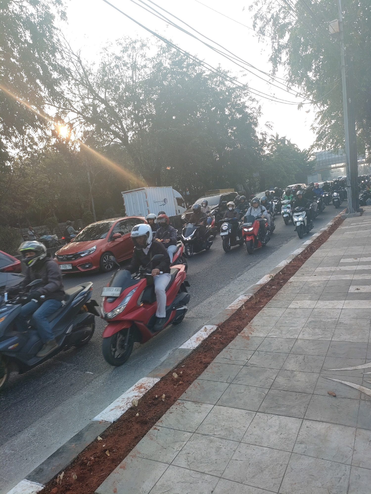 The image depicts a busy street scene in Bekasi, Indonesia, likely during the morning commute. A long line of motorcycles and a few cars are moving along the road, indicating moderate traffic. The sun is low in the sky, casting a warm glow and lens flare across the image, suggesting it is either sunrise or sunset. The road is lined with trees on the left and a sidewalk with patterned tiles on the right. Some construction or landscaping work is evident along the edge of the sidewalk, with exposed soil and young plants. The overall atmosphere is one of everyday urban life in Indonesia, with a focus on the prevalent use of motorcycles for transportation.