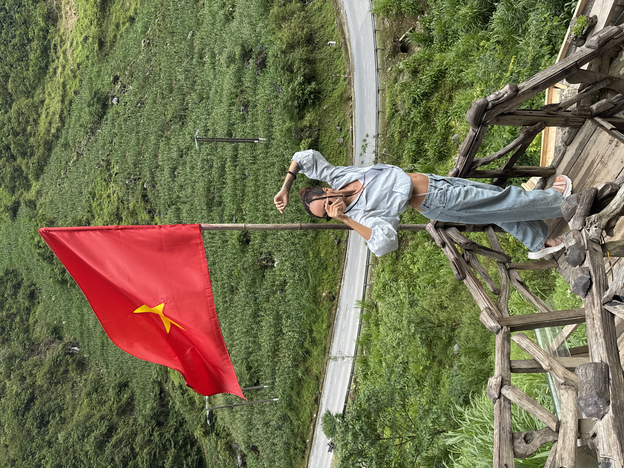 A medium shot reveals a person standing on a wooden structure overlooking a landscape in Yên Minh Commune, Vietnam. The individual, wearing a light blue top, jeans, and sandals, is captured in a contemplative pose with sunglasses perched on their nose. A red Vietnamese flag with a yellow star is prominently displayed nearby. The environment is lush and green, with a road winding through the landscape. The wooden structure the person is standing on appears to be part of a viewpoint or lookout point.