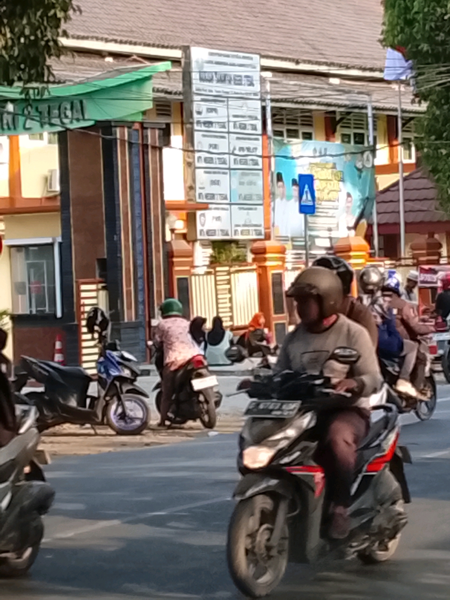 The image depicts a street scene in Slawi, Indonesia, likely in front of a government or educational building, suggested by signage and architectural elements. In the foreground, several people are riding motorcycles. The motorcycle closest to the viewer has a rider wearing a beige long-sleeved shirt and dark pants, with a helmet covering their head. Another motorcycle behind it has two riders, the passenger wearing a white headscarf and the driver wearing a brown jacket and helmet. A third motorcycle is partially visible to the left. Further back, several individuals are seated on the side of the road, some wearing headscarves. In the background, a building with a tiled roof and several windows is visible. Large signs, one green with text and another with multiple columns of text, are mounted on poles in front of the building. A banner with images of people and text is also displayed. The street is paved and appears to be a relatively busy thoroughfare. Trees are visible on the right side of the image.