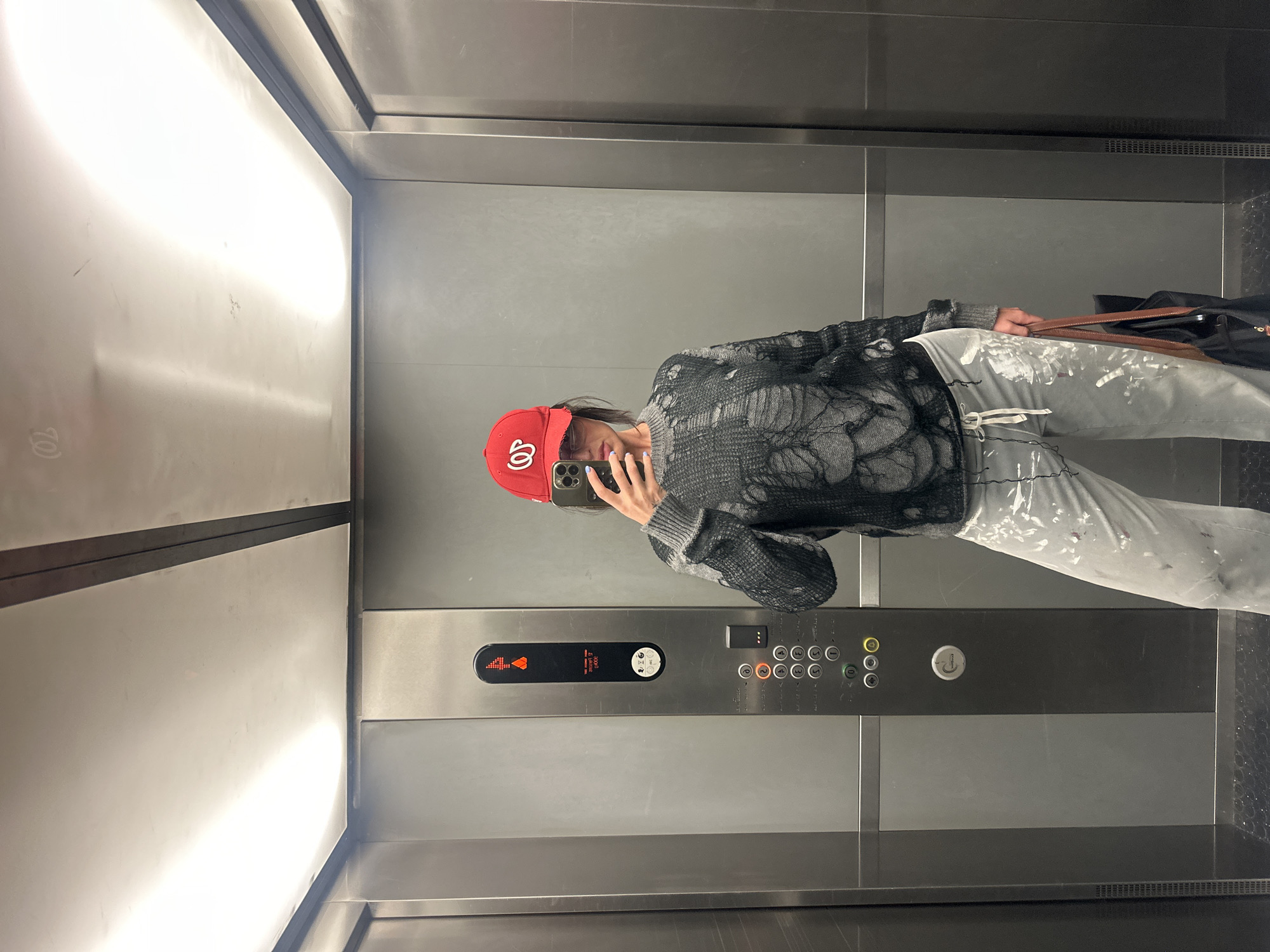 A person wearing a red baseball cap, a grey sweater with a distressed design, and grey pants with white paint splatters, is taking a selfie in an elevator. The elevator is made of stainless steel and has a panel with floor buttons, as well as illuminated indicators showing the current floor as "4" and the direction of movement as "up". The person is holding a black smartphone in their left hand to take the picture. A black bag with brown leather straps is visible in the upper right corner of the frame. The location is unknown, and there is no visual information to suggest it is in London, United Kingdom.