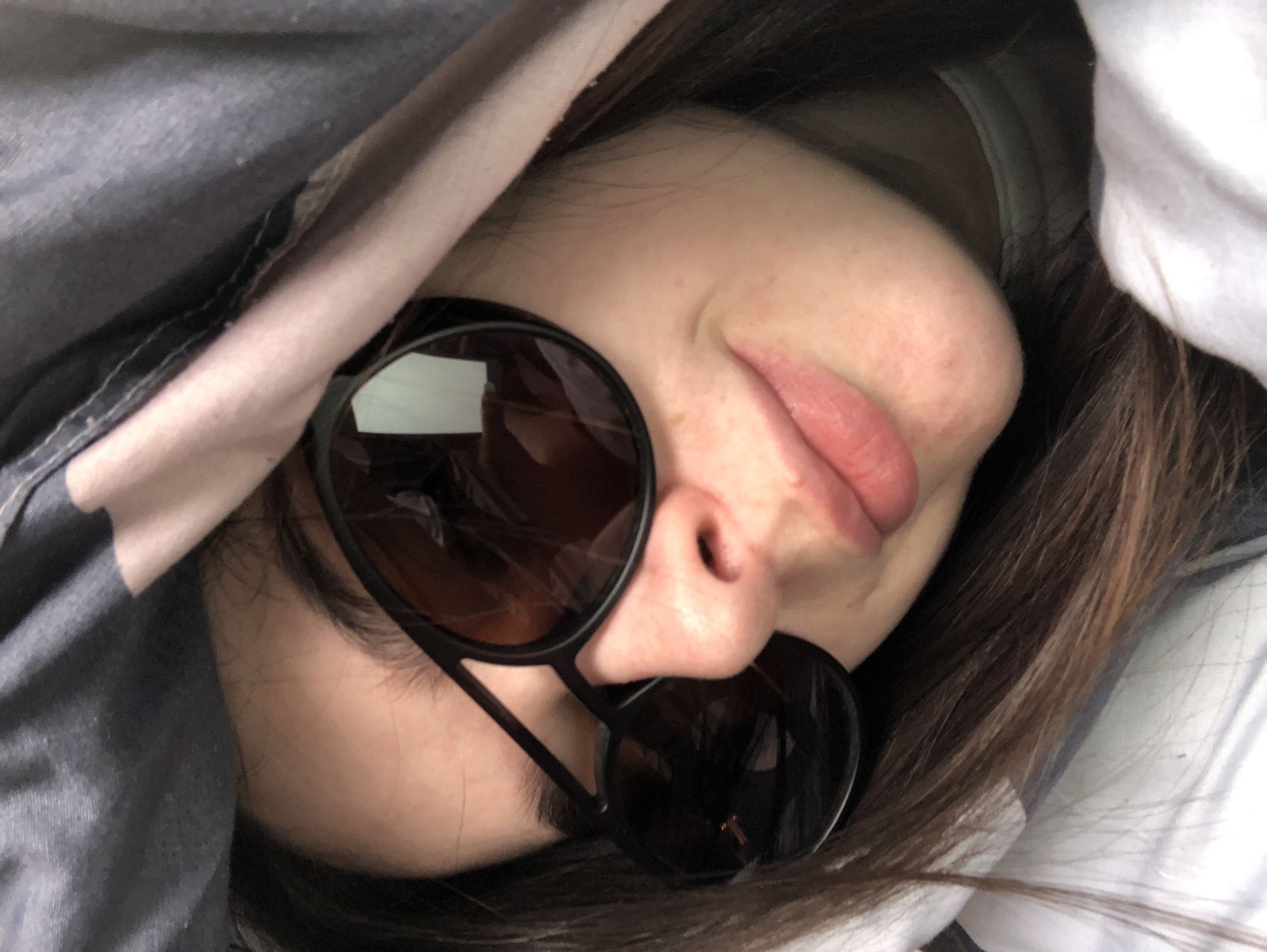 A close-up, slightly angled photograph depicts a person with dark hair partially covered by a grey patterned duvet and a white pillow. The individual is wearing round-framed sunglasses. The background is mostly obscured by the bedding. The location is unknown; there is no indication that the photograph was taken in Chaikovsky, Russia.