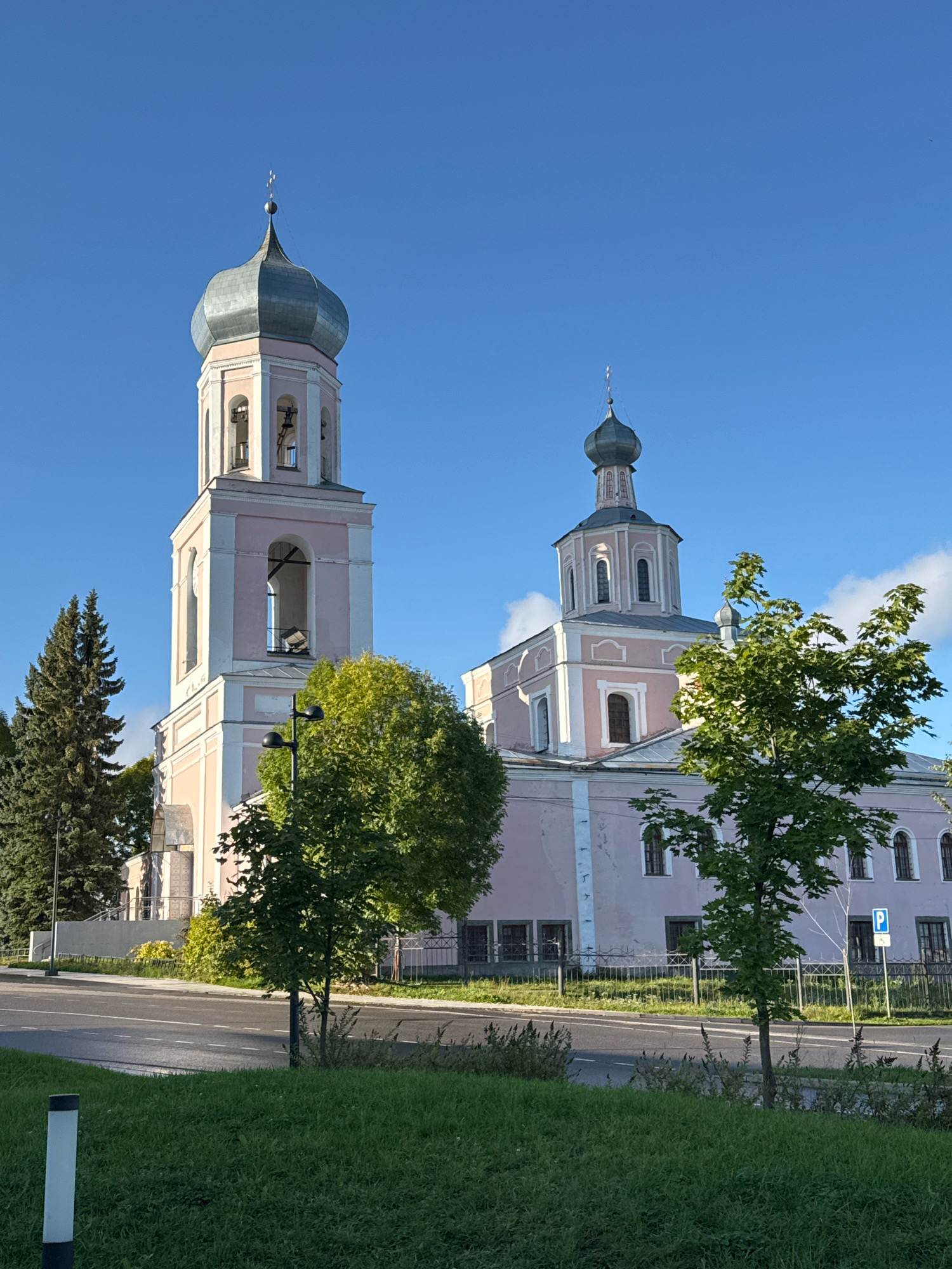 The image shows the bell tower and main building of the Resurrection Cathedral in Valday, Russia, on a clear day with a bright blue sky and a few wispy clouds. The cathedral is painted a light pink with white trim. The bell tower is tall and square at the base, tapering to an octagonal section topped with a silver dome. The main building is rectangular and has a smaller, similarly shaped dome. Several mature green trees are in the foreground and to the left of the cathedral. A paved road runs in front of the building, separated from the cathedral grounds by a black metal fence. Green grass covers the foreground. A parking sign with a blue "P" is visible on the right side of the image, behind the fence. No people are visible in the frame.