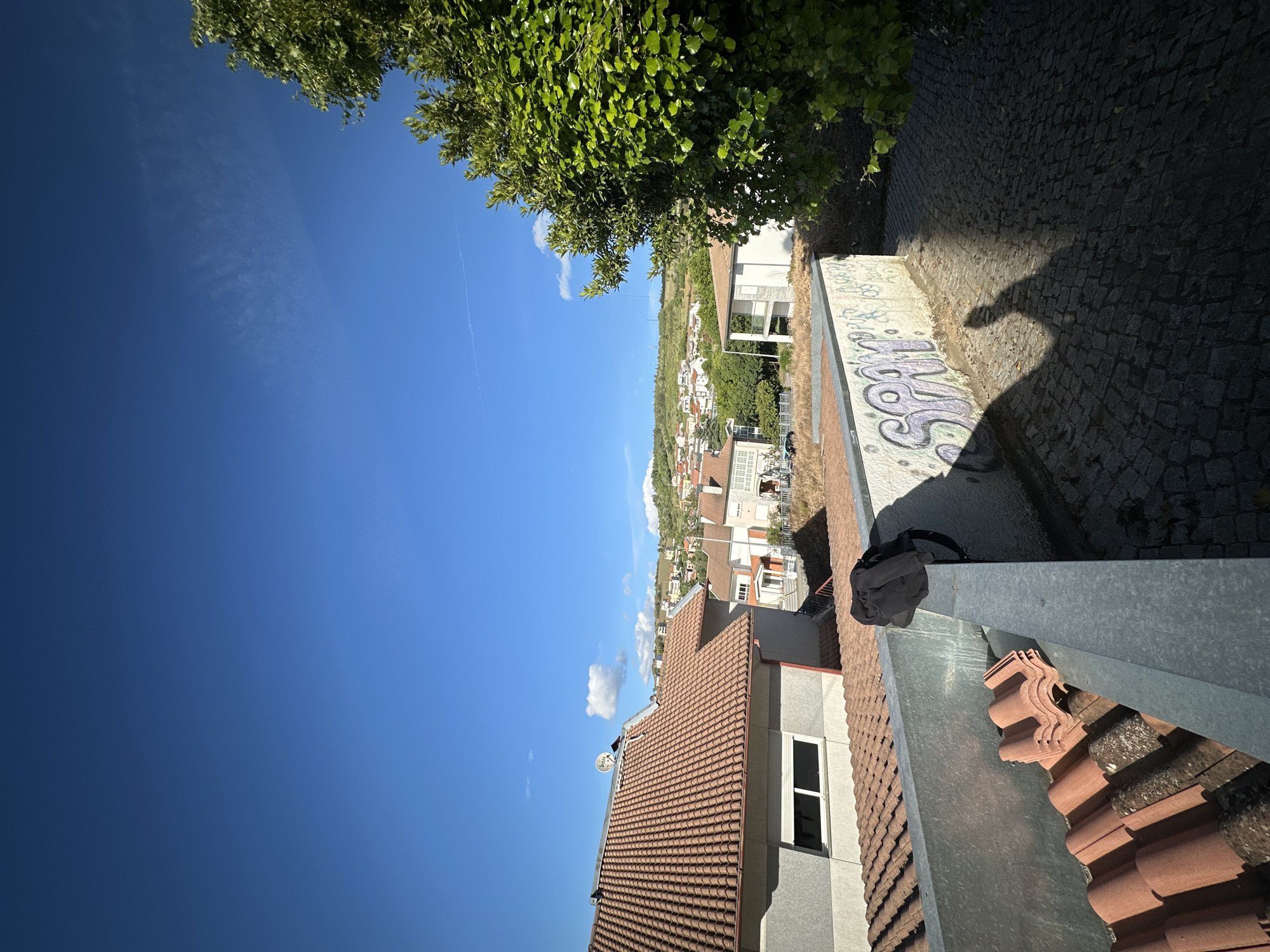 A high-angle, outdoor shot from a rooftop in Bragança, Portugal, shows a clear blue sky and a townscape. At the top left, a tree is in frame. The lower part shows a tile roof with a window. A grey metal edge is visible. In the background, there are houses and a road with graffiti. The shadow of a person is visible on the road. A bag is on the metal edge. The image is taken during daytime.