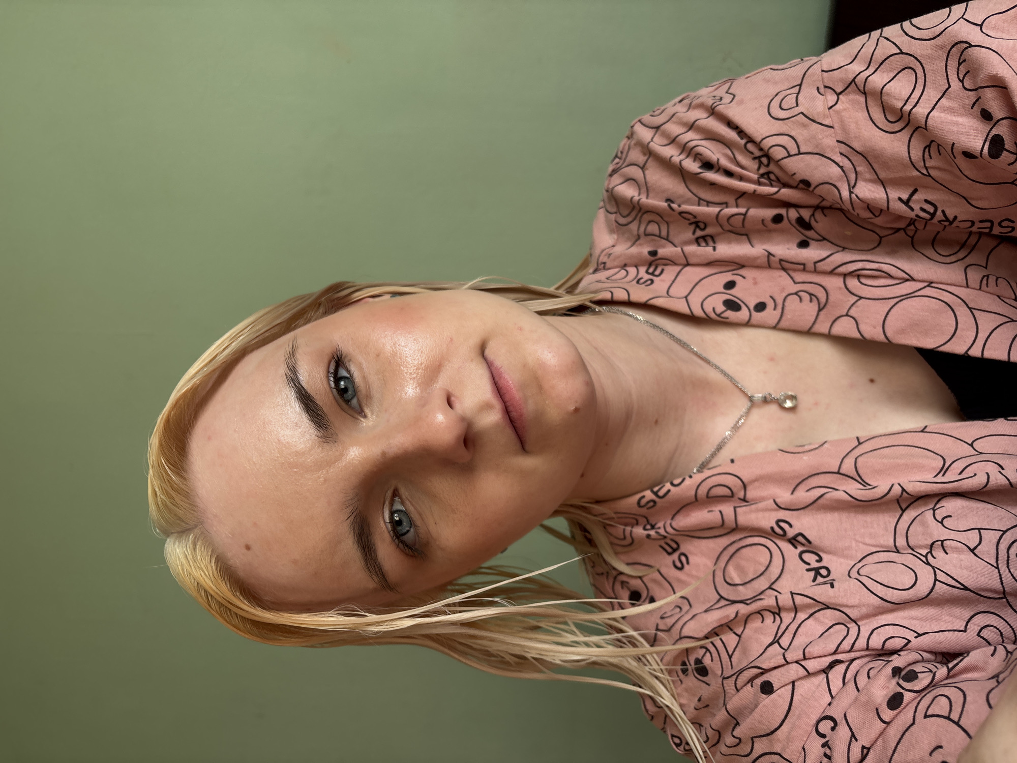 A close-up, rotated portrait shows a young woman with blonde hair and blue eyes, looking directly at the camera with a subtle, calm expression. Her face is slightly angled, and her blonde hair, which appears somewhat damp, frames her face and shoulders. She has dark, well-defined eyebrows that contrast with her light hair.
She is wearing a light pink top, possibly a casual shirt or pajama top, featuring a repeating pattern of outlined, cartoon-style bear figures and the word "SECRET" in black text. A delicate silver chain necklace with a small, clear pendant hangs around her neck. The background is a plain, muted green wall, indicating an indoor setting. The image is well-lit, highlighting the sheen on her skin and the details of her clothing. The text "SECRET" is clearly visible on her shirt multiple times.