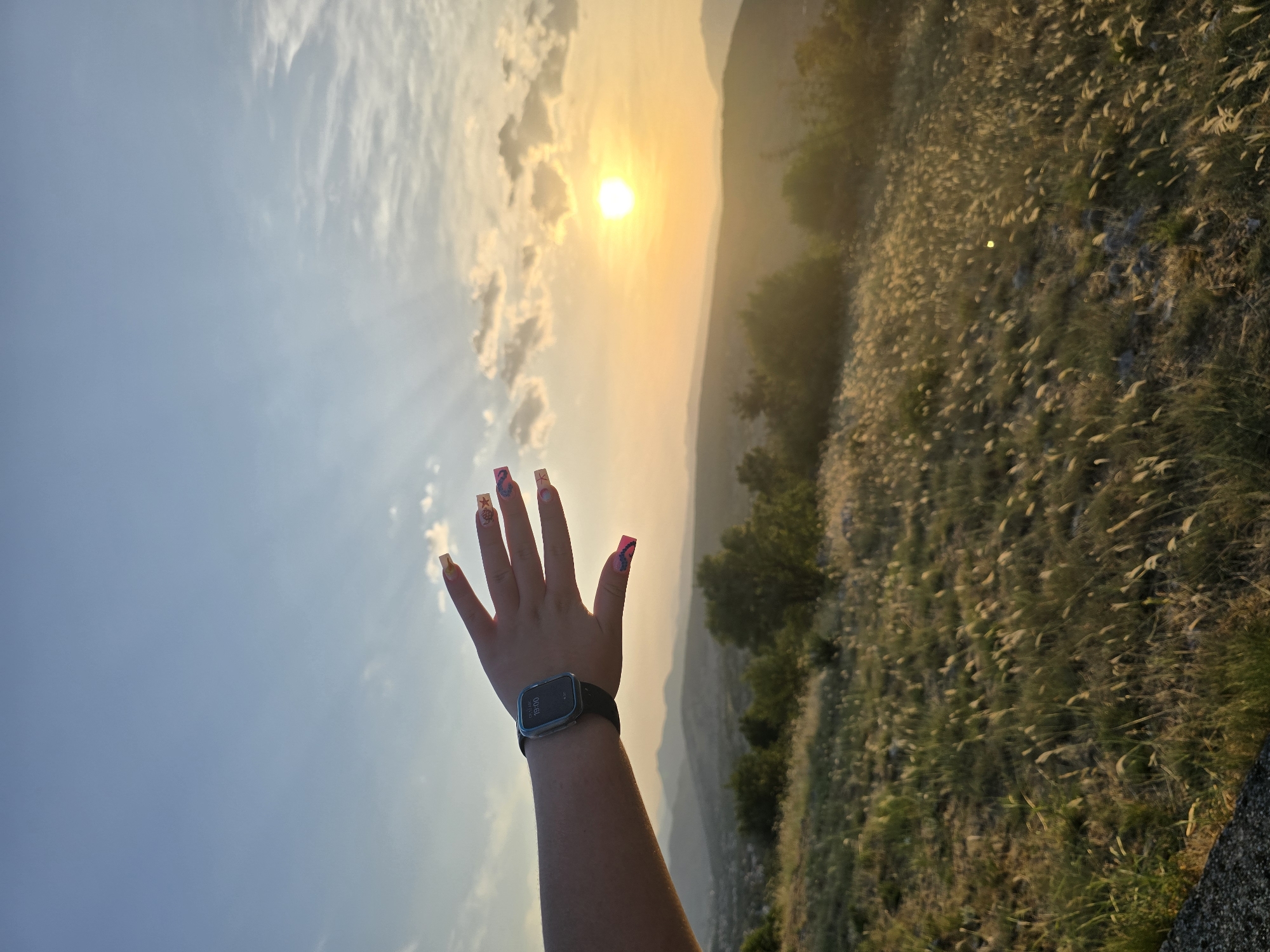 An over-the-shoulder, eye-level shot captures a person's left arm and hand extended towards the setting sun. The person is wearing a smartwatch on their wrist, displaying the time "19:00." The hand is open, with fingernails painted in bright colors and adorned with decorative designs. The background features a panoramic view of rolling hills and mountains bathed in the warm, golden light of the sunset. A vast expanse of dry grass and sparse vegetation covers the foreground and midground. The sky above is a gradient of blue and orange, with scattered clouds catching the last rays of sunlight. The scene appears to be an outdoor location, likely on a hilltop or elevated area, with a settlement visible in the distance among the hills. While the exact location in Podgorica, Montenegro cannot be definitively determined from the image alone, the landscape suggests a natural setting on the outskirts of the city.