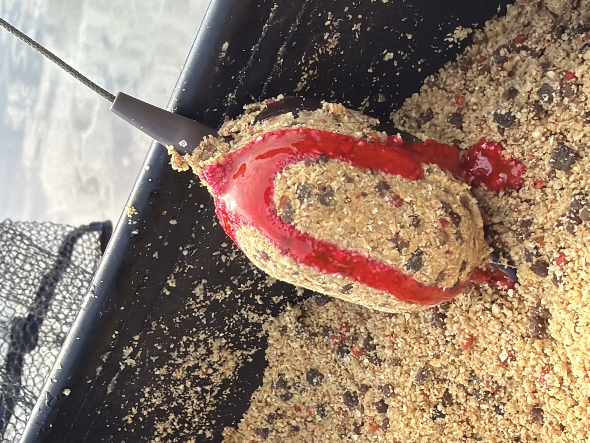 The image shows a fishing feeder, a type of weight and bait container used in angling, submerged in a mixture of groundbait. The feeder is partially covered with a red, viscous liquid, possibly an attractant or flavor enhancer. The groundbait appears to be a blend of particles, likely including cereal-based ingredients, seeds, and other components used to attract fish. The feeder sits inside a black container, with a fishing net partially visible in the background on the left side. The specific location within Prikamskii, Russia, is not discernible from the image. There are no people visible in the image.