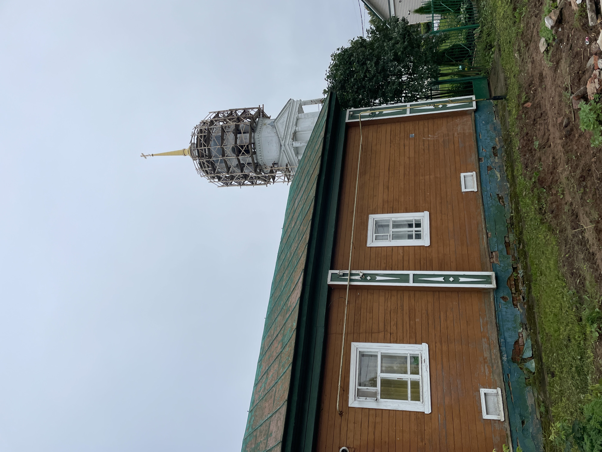 In the city of Kstynino, Russia, a wooden house with a weathered green roof stands in the foreground. The house is painted brown and features white-trimmed windows. The foundation of the house is visible, showing exposed brickwork and peeling green paint. To the right of the house, a tall white structure, possibly a church bell tower, is partially obscured by scaffolding. A golden cross tops the tower. A mature tree with dark green foliage grows next to the tower, and a green metal fence encloses a garden area with a white house in the background. The sky is overcast, casting a muted light over the scene. The ground in front of the house is uneven, with patches of grass and disturbed soil, suggesting recent construction or landscaping work.