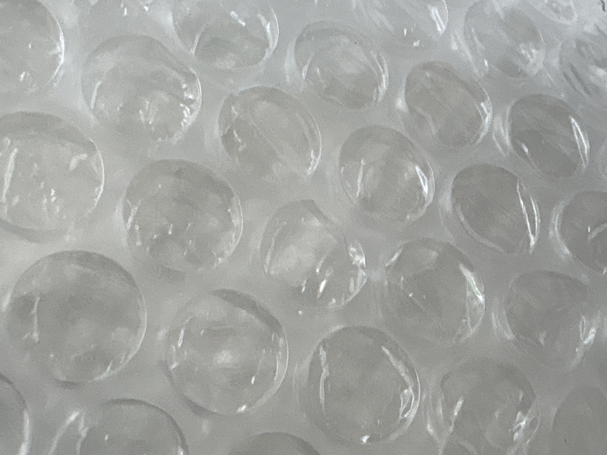 The image displays a close-up, high-angle view of transparent bubble wrap. The primary subject is the textured surface of the protective packaging material, characterized by numerous individual air-filled cells arranged in a repeating pattern. These bubbles are translucent, allowing for a diffused view of a light, possibly white or light grey, surface beneath. Each bubble exhibits subtle reflections and distortions on its rounded surface, indicating an ambient light source.
There are no people or any discernible activities taking place in the image. It is a static depiction of an object. No specific time of day or weather conditions can be determined from the uniform, soft lighting. There are no direct location cues within the image itself to suggest Saint Petersburg, Russia, as provided in the context. No emotions are conveyed by the inanimate subject, and no visible text appears in the frame. The overall impression is one of texture and repetitive form.