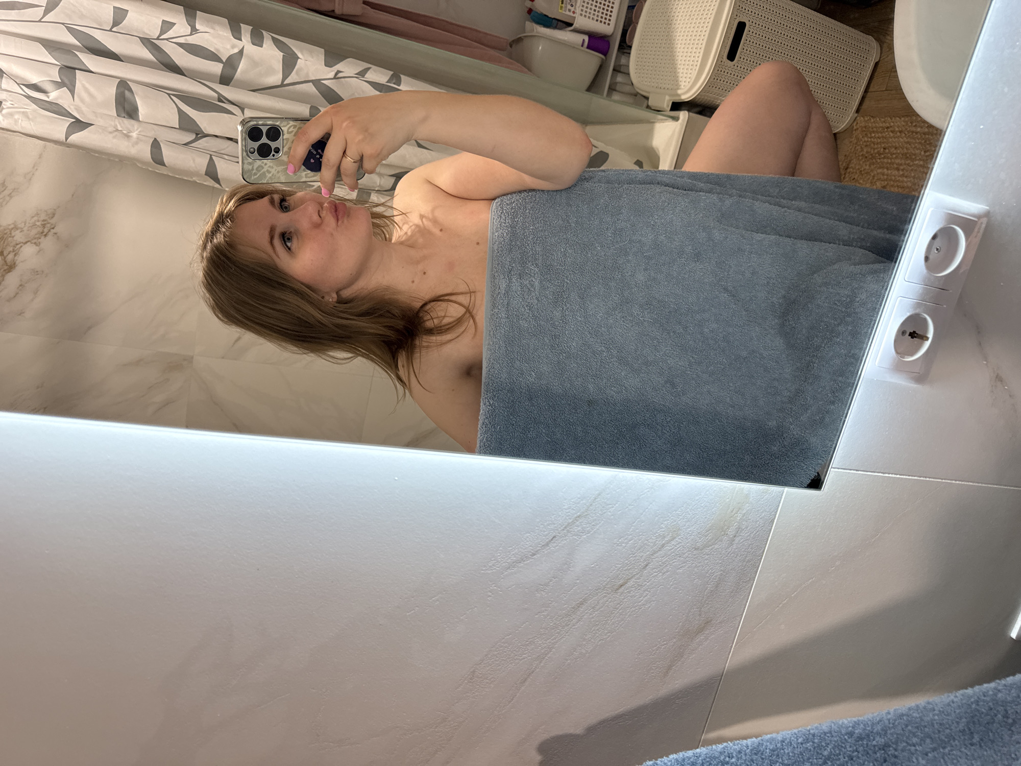 A woman is in a bathroom, taking a selfie in a mirror. She is partially covered by a light blue towel. Her face is visible, as she holds up a smartphone with her left hand. Behind her, there is a shower curtain with a leaf pattern. To the right in the background, there are white storage containers. The bathroom has light-colored tiles on the walls and floor. An electrical outlet is visible on the wall near the mirror. The location is Ферма, Russia.