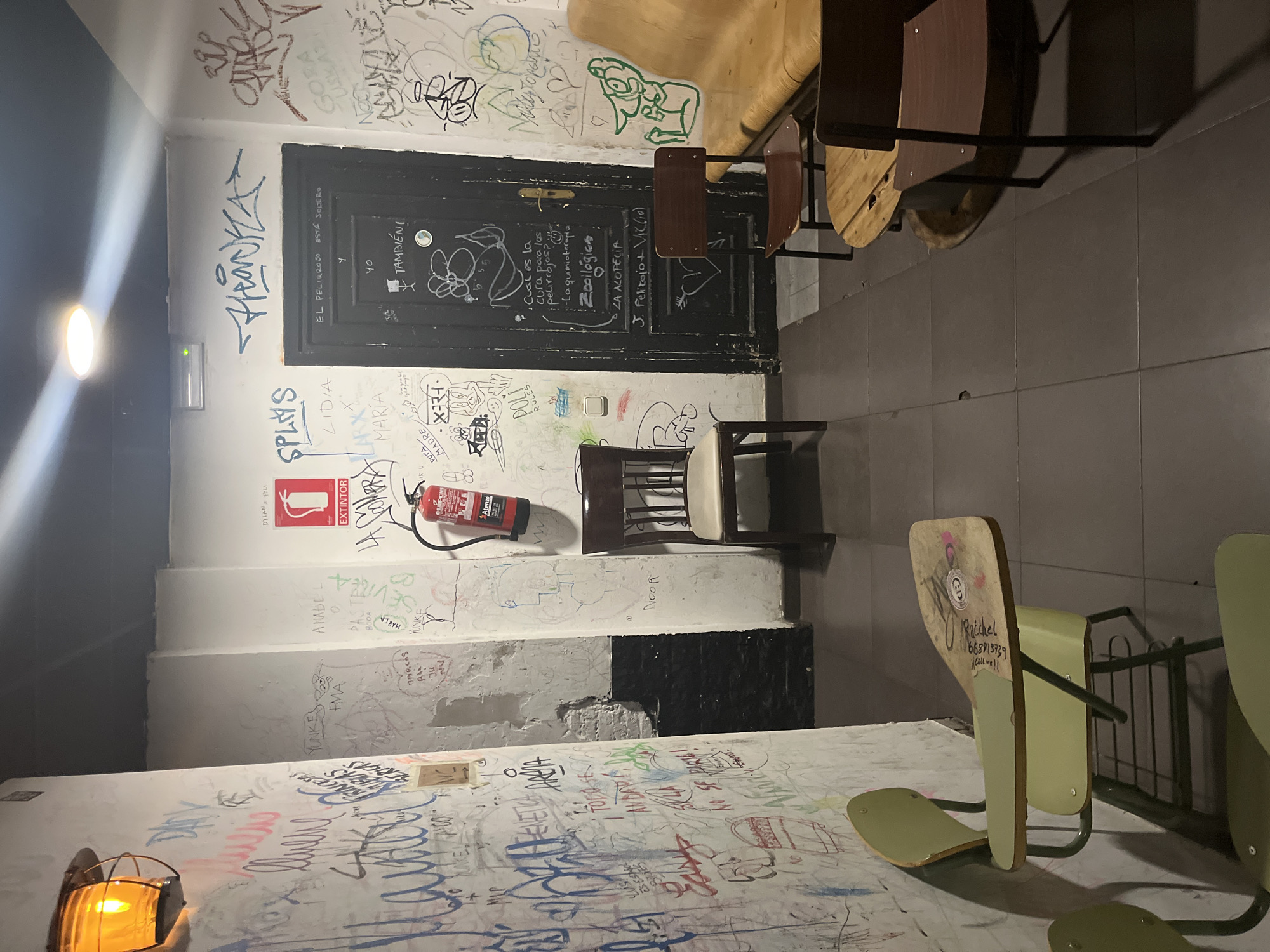 The image captures an informal indoor space in Valencia, Spain, characterized by heavily graffitied walls and a dark, worn aesthetic. No people are visible. The scene is dominated by white walls completely covered in an eclectic mix of handwritten messages, tags, doodles, and graffiti in various colors, suggesting a long history of visitor interaction.
A prominent feature is a dark wooden door, also covered in chalk and pen marks, situated in the center-left. Next to the door, a red fire extinguisher is mounted on the wall. Several chairs are present; two light brown wooden chairs with dark metal frames are positioned near the door, and additional school-style chairs with light green seats and backs are seen towards the bottom right, some stacked or leaning. An orange-tinted industrial-style light fixture illuminates the bottom-left corner, and another bright overhead light source casts a strong glare on the left side of the image. The floor consists of dark grey tiles.
The activity implied is the leaving of personal marks and messages by visitors over time. Notable textual details from the graffiti include: "SPLAYS," "THANKA," "EL PELIRROJO ESTA SOLTERO" (The redhead is single), "TAMBIÉN!" (Also!), "Cual es la cura para los pelirrojos? -La quimioterapia" (What is the cure for redheads? -Chemotherapy), "Zoollogies," "LA ALOPECIA" (Alopecia), "J. Pelizojo + VIC(10)," "DYLAN POLI," "ANABEL," "LIDIA," "MARIA," and "Raichel 668813739." The fire extinguisher is labeled "EXTINTOR." The setting appears to be a late-night or artificially lit venue, like a bar or a social club, given the internal lighting and the nature of the wall markings.