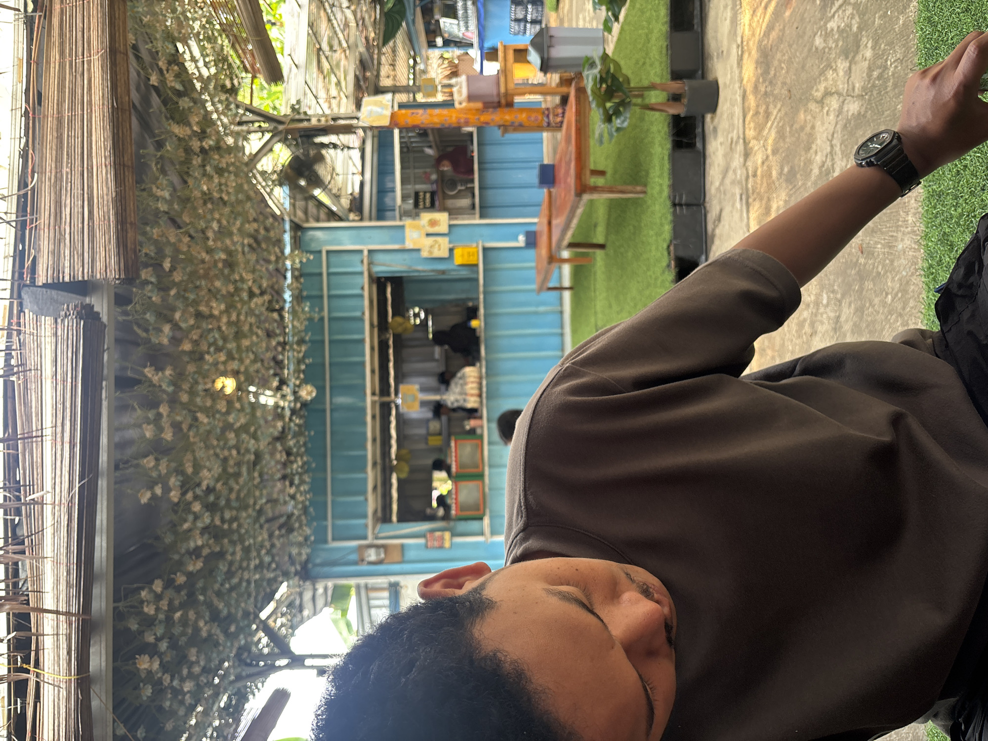 An overhead shot shows a man with dark curly hair, a mustache, and a dark grey t-shirt sitting at a table in an outdoor cafe in Tangerang, Indonesia. The cafe has a rustic, natural aesthetic with a thatched roof covered in dried flowers. In the background, blue corrugated metal structures house food stalls. Green artificial grass covers the ground, interspersed with wooden tables and benches. A black watch is visible on the man's left wrist. The lighting suggests it is daytime.