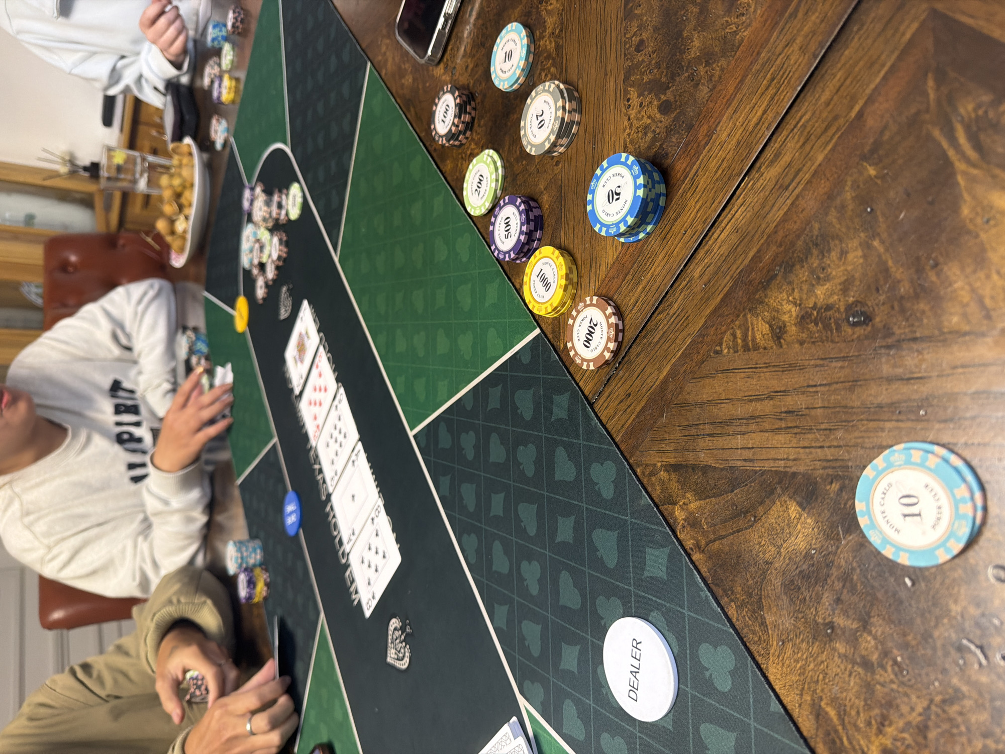 The image shows a group of people playing poker around a table. The table has a green felt playing surface with a Texas Hold'em diagram printed on it. There are several stacks of poker chips on the table, in various denominations and colors. Some cards are also visible on the table, including what appears to be the flop of a hand. One person's hand is holding a few chips, suggesting they are actively participating in the game. Another person has their hands resting on the table near the cards. The environment appears to be an indoor setting, possibly a dining room or living area. The background shows a portion of a wooden table and chairs, and a sideboard with a glass of water and a plate of food. There is no discernible information about the specific location within Xuan Huong - Da Lat, Vietnam.
