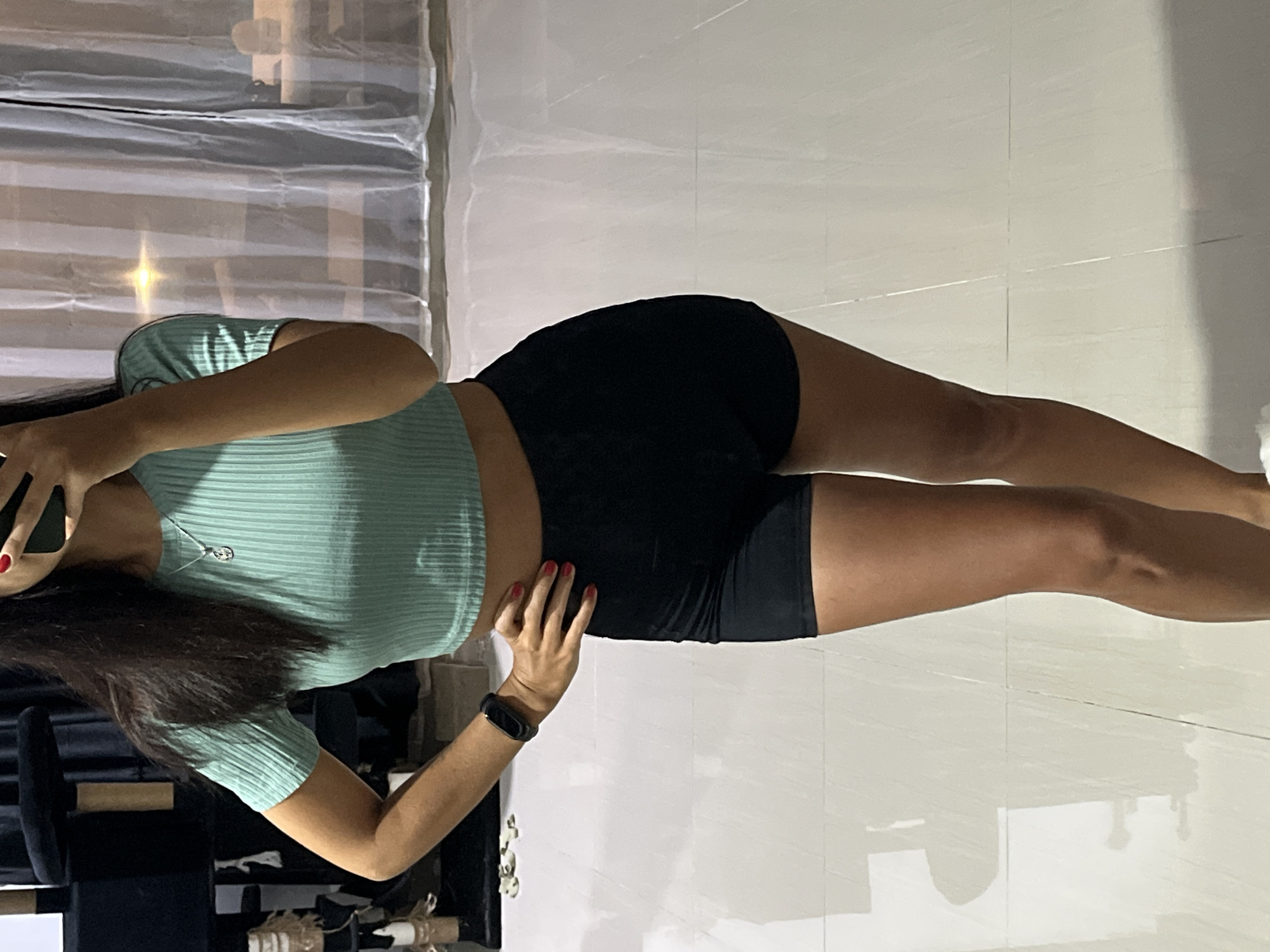 A woman is in what appears to be an indoor setting. The backdrop includes what looks like a partially visible curtain and other furniture. The woman has dark hair and is wearing a light green short-sleeved top and dark-colored shorts. Her left arm is extended and slightly bent while holding a phone to her face, while her right hand is resting near her right hip. There is a fitness tracker on her right wrist. The floor is a light-colored tile. The environment does not have distinct landmarks to identify that it is Kuta Selatan, Indonesia.